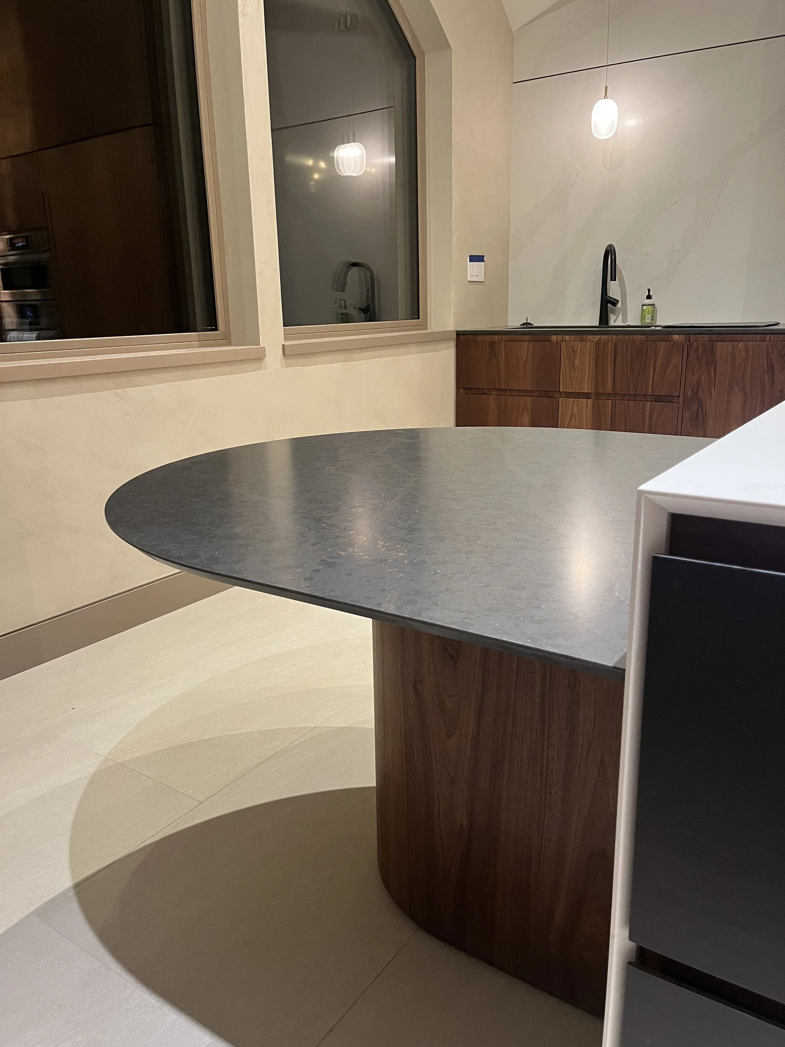 A modern kitchen with a black countertop, wooden cabinets, and a black faucet, seen through a large window. There are two hanging pendant lights and a soap dispenser on the counter.