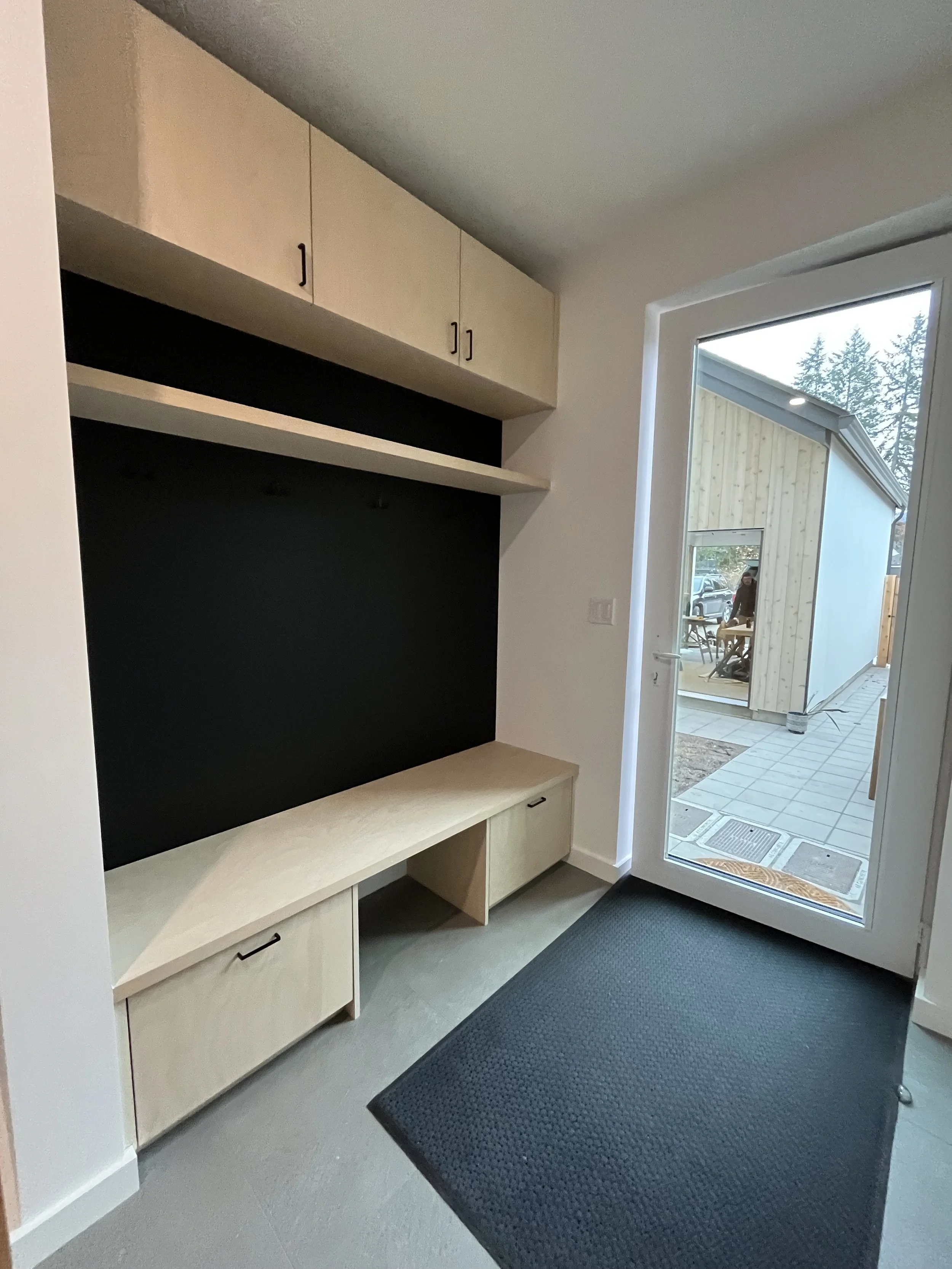 Interior view of a mudroom with a bench, upper and lower cabinets, a black accent wall, and a glass door leading outside to a patio area.