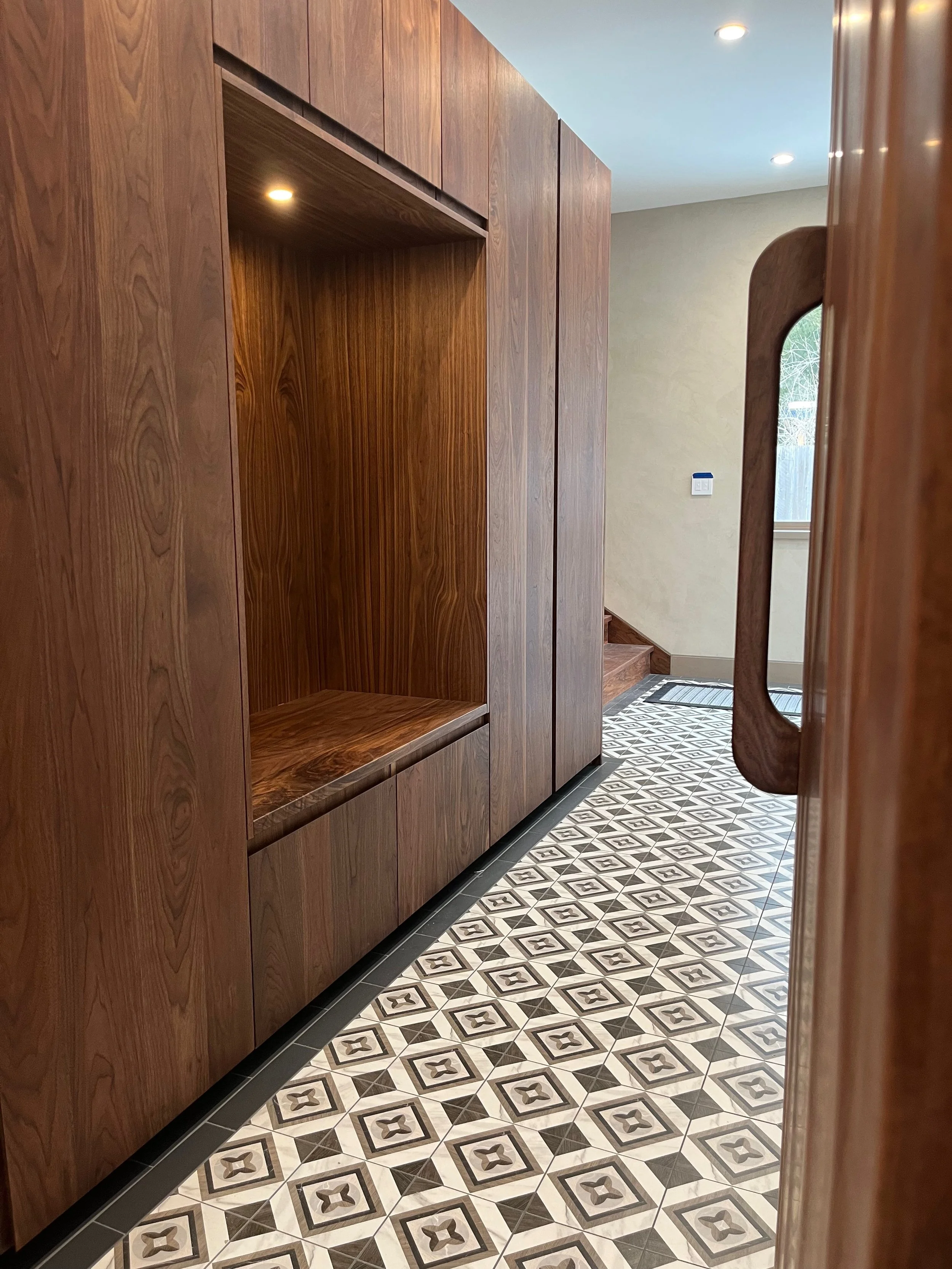 View of a hallway with patterned tile floor, wooden cabinets and stairs in the background, and a window letting in natural light.
