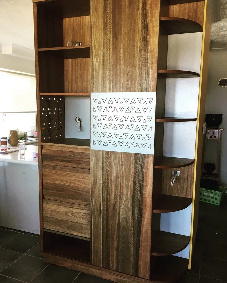 A wooden shelving unit with multiple open shelves and drawers, decorated with a white geometric patterned panel in the center.