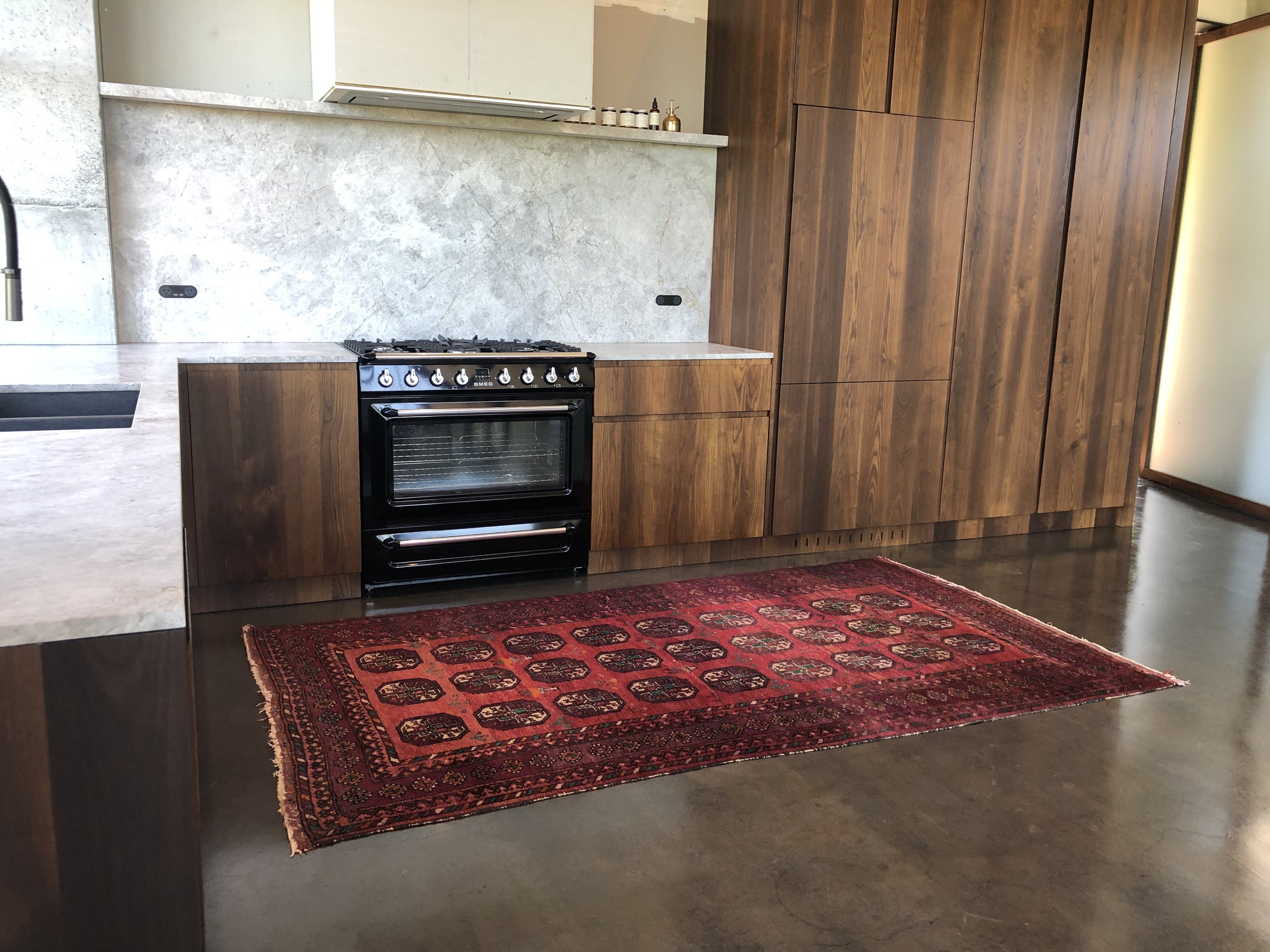 Modern kitchen with wooden cabinets, black oven, gray countertop, and red patterned rug on polished concrete floor.