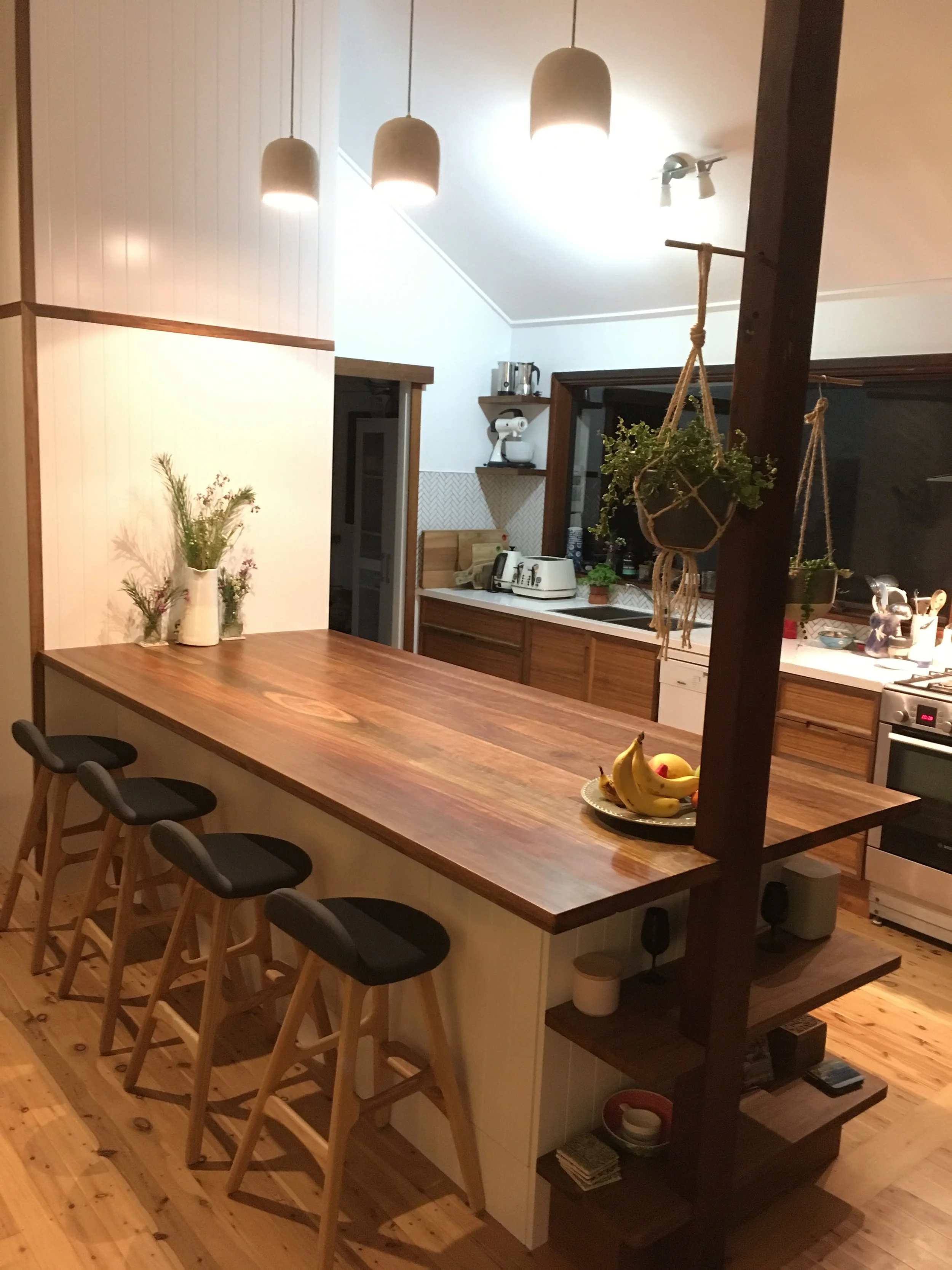 A wooden kitchen island with four black and wooden stools, decorated with a bowl of bananas and apples, and plants hanging from a wooden post. In the background, a kitchen with white and wooden cabinets, a countertop with small appliances, and a wind