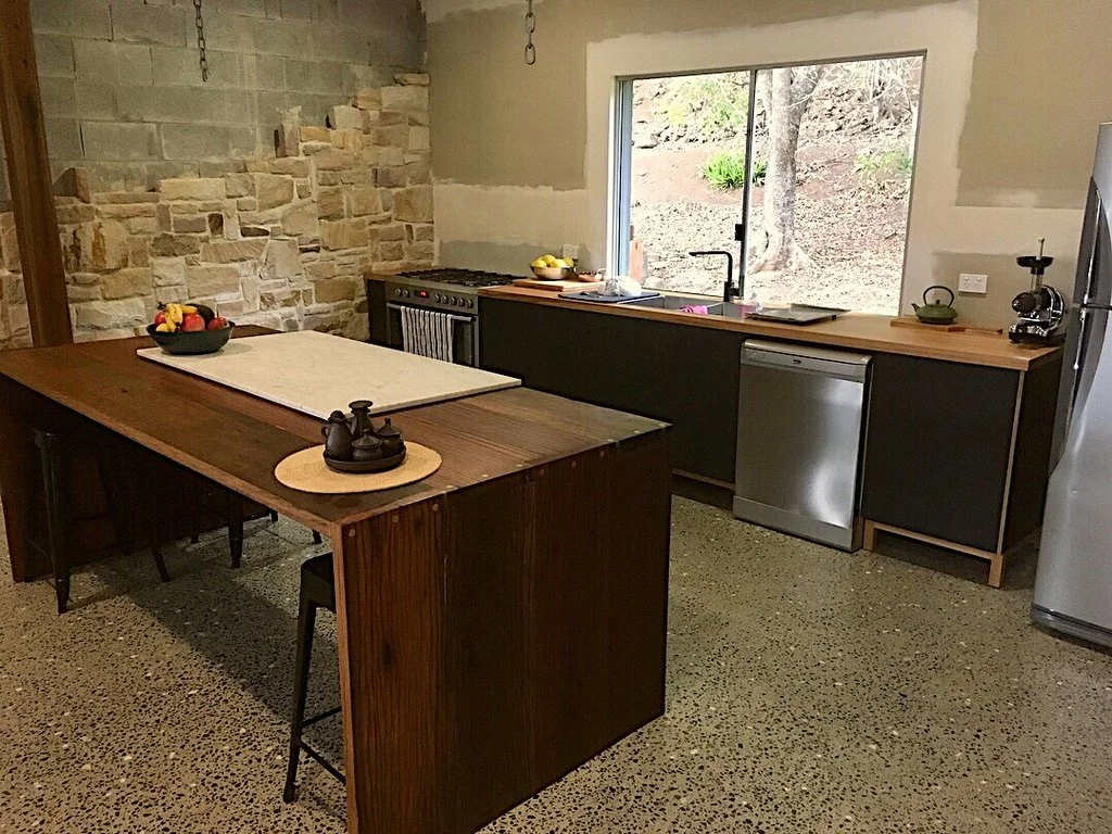 A modern kitchen with a large wooden island, black and wooden cabinets, a stainless steel dishwasher, a window overlooking a dirt yard, and a stone accent wall.