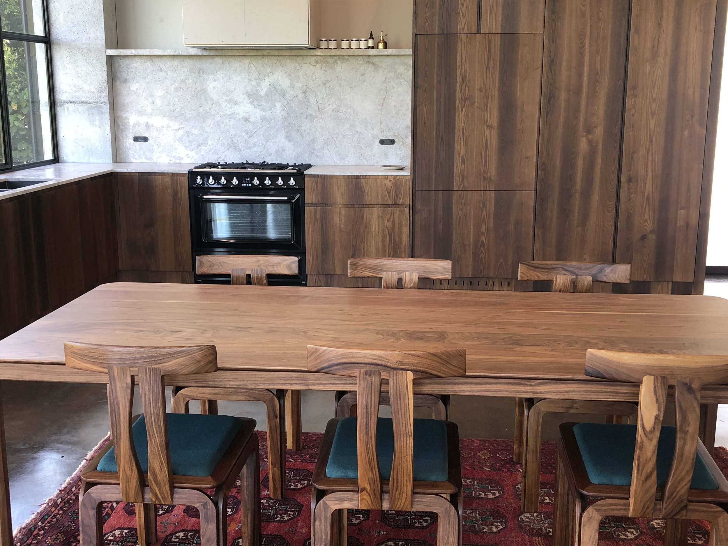 A modern kitchen with wooden cabinets, a black stove, and a wooden dining table with six chairs. The setting includes a Persian rug under the table.