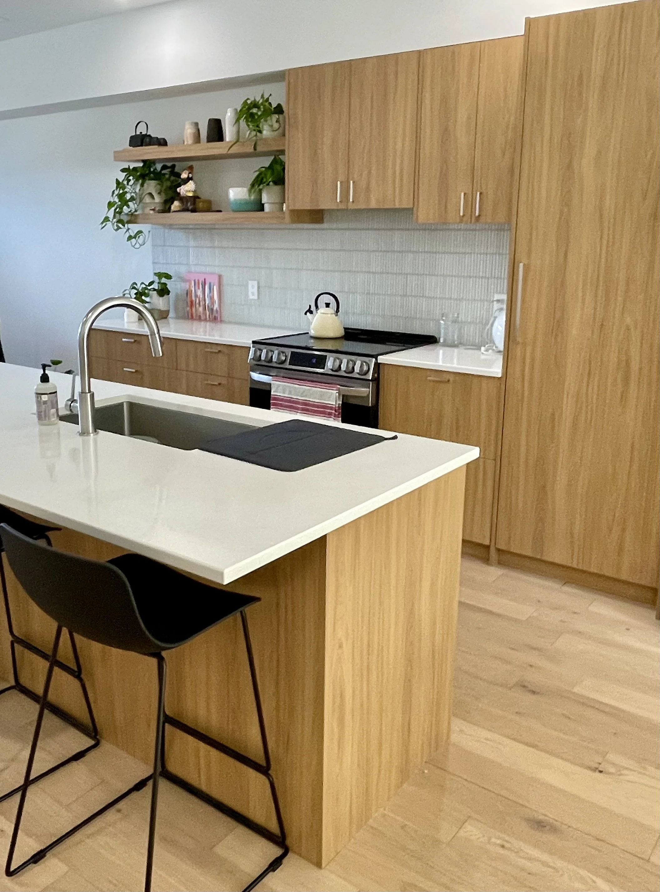 Modern kitchen with wooden cabinets, white countertop island, black chairs, and decorative plants on open shelves.