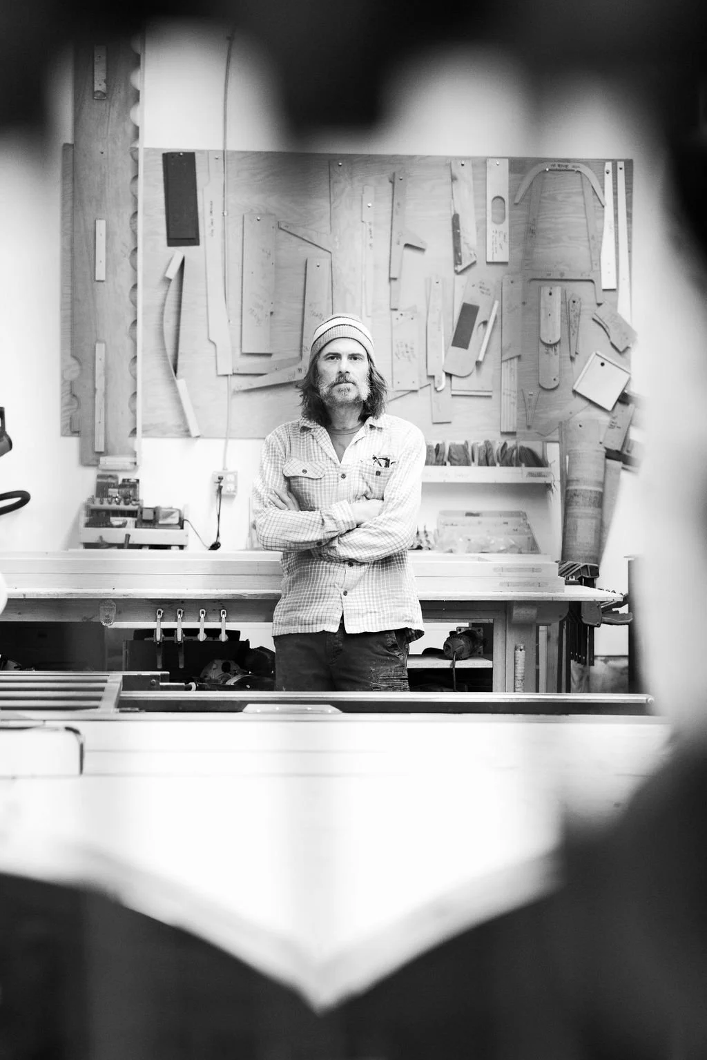 A man with long hair and a beard, wearing a striped beanie and a checkered shirt, standing with his arms crossed in a woodworking workshop. The workshop has wooden boards and tools on the wall behind him.