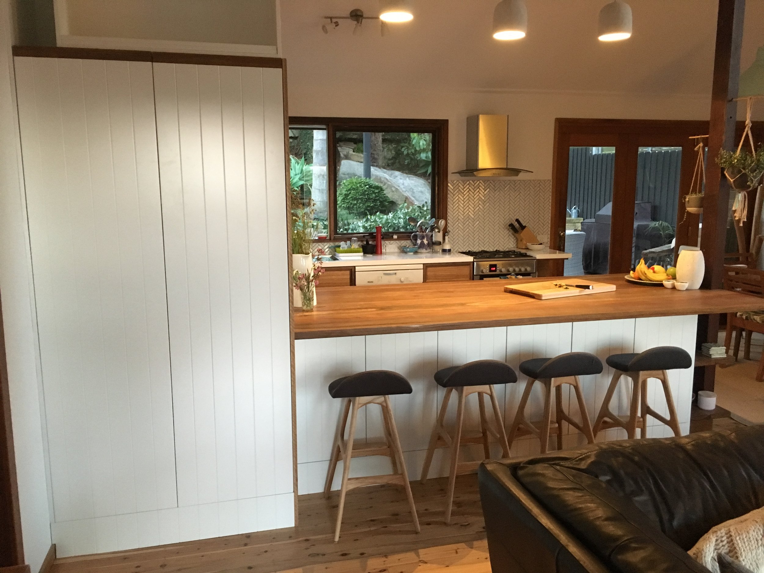 Kitchen with wooden countertop, four black bar stools, stove, window with outdoor greenery, sliding glass door, hanging pendant lights, and various kitchen items.