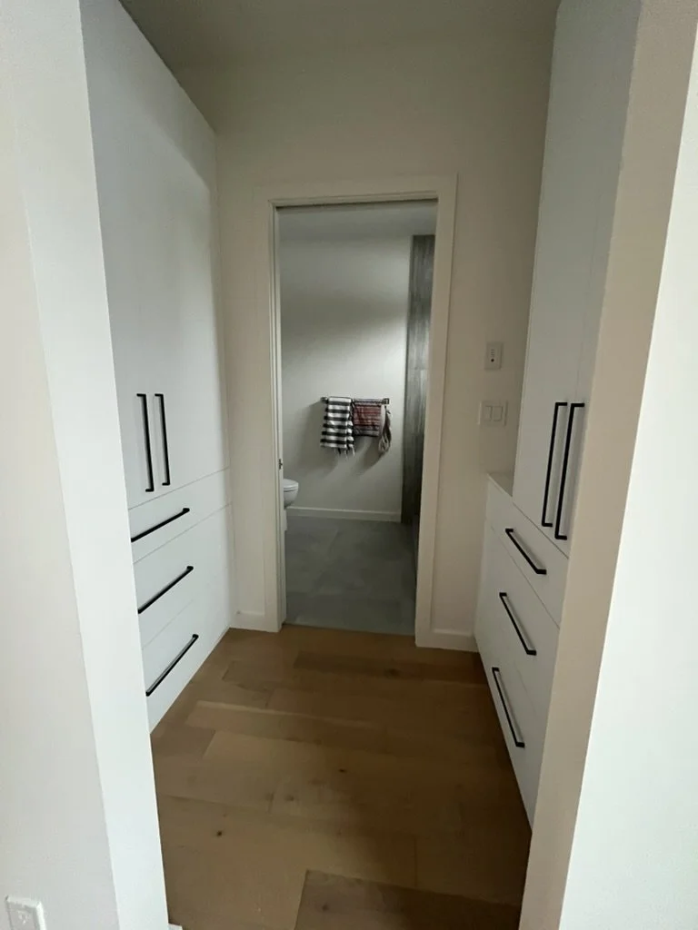 Interior view of a hallway leading to a bathroom with towels hanging on a towel rack. White cabinets with black handles are visible on both sides of the hallway. Wood flooring in the foreground and tile in the bathroom.