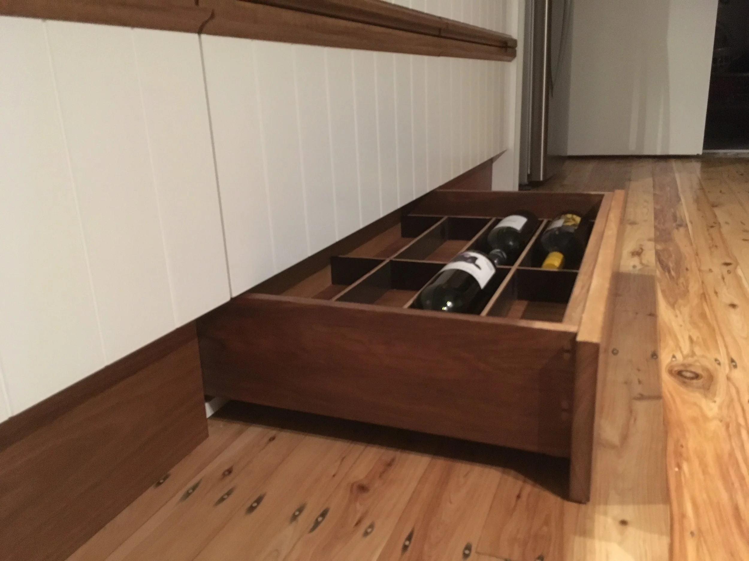 A wooden pull-out wine rack stored under a white kitchen cabinet, holding several wine bottles, located on awooden floor in a kitchen.