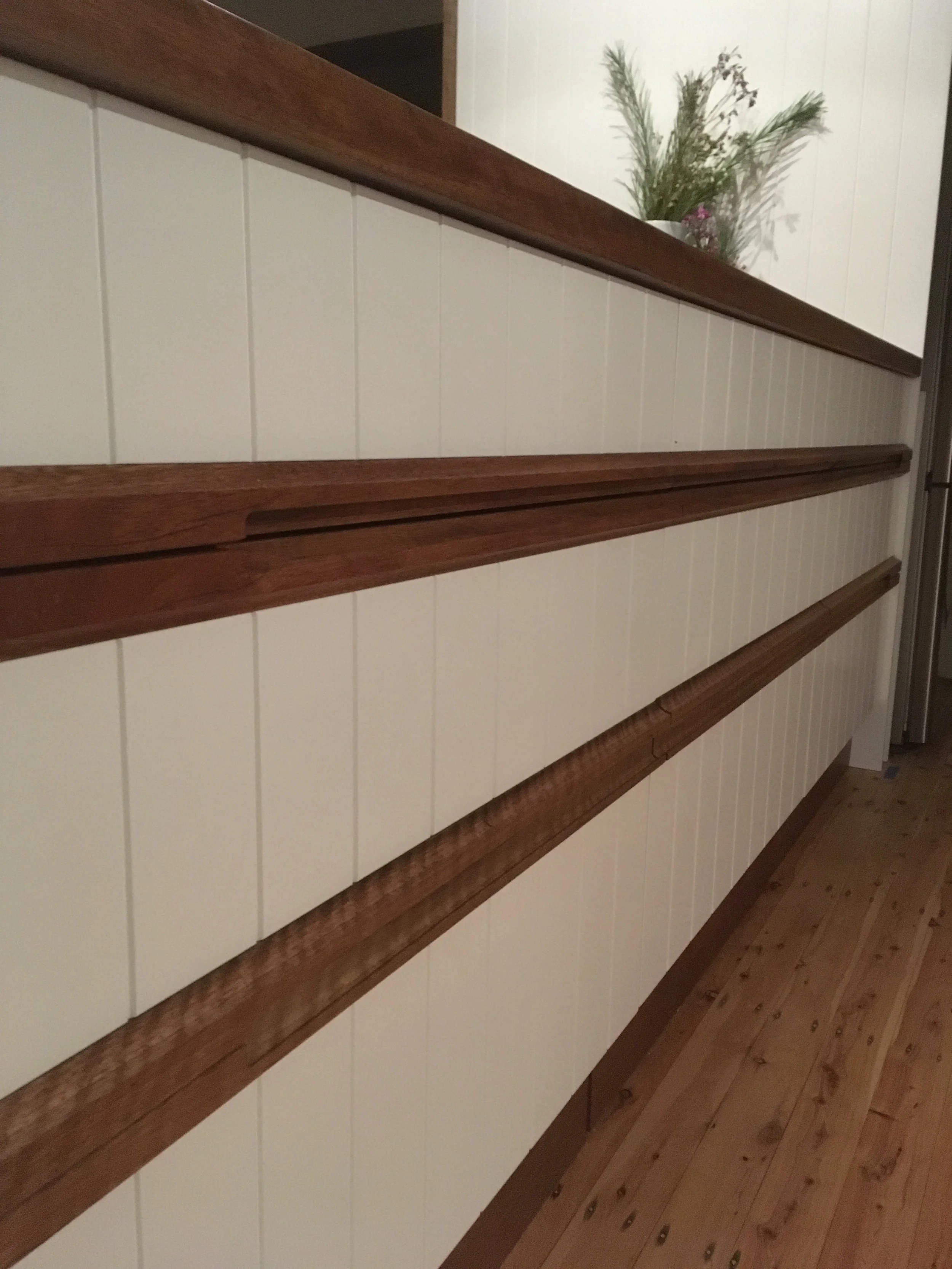 A wooden staircase with white paneling on the side, topped with a wooden handrail. There is a plant with green foliage and purple flowers at the top of the staircase.