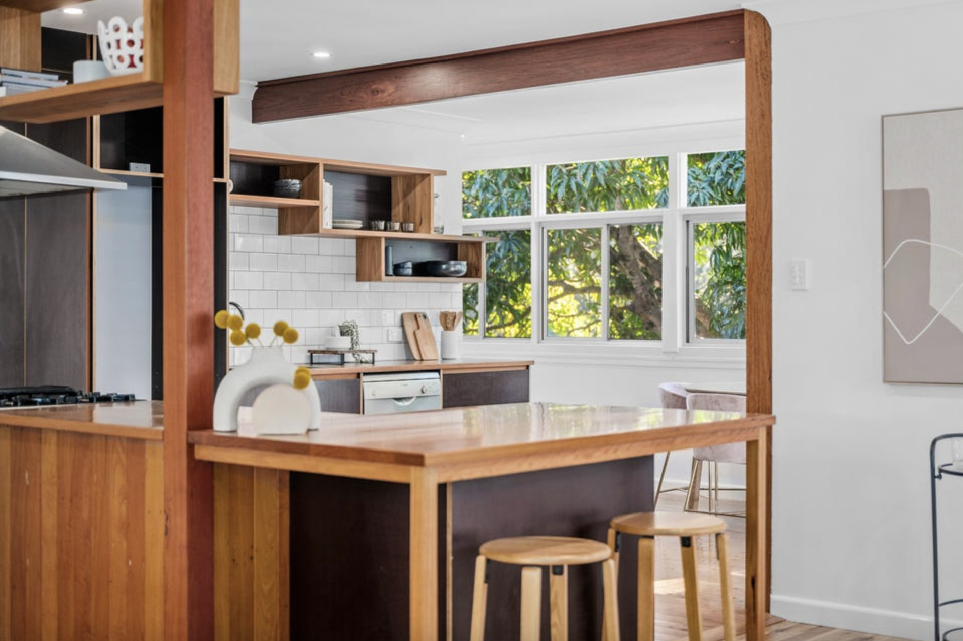 Bright kitchen with white subway tile backsplash, wooden open shelves, and large windows showing lush greenery outside.