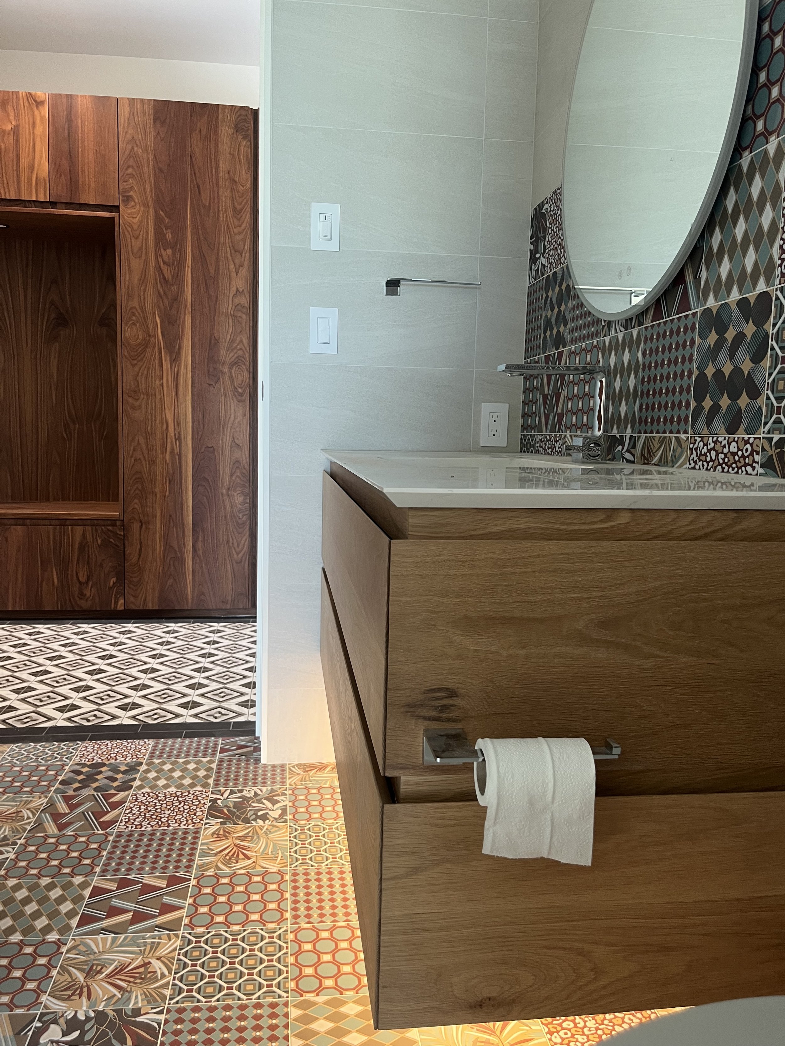 Bathroom with a wooden cabinet, a patterned tile floor, a mirror, a sink with a faucet, a paper towel holder with a roll of paper towel, and light-colored wall tiles.