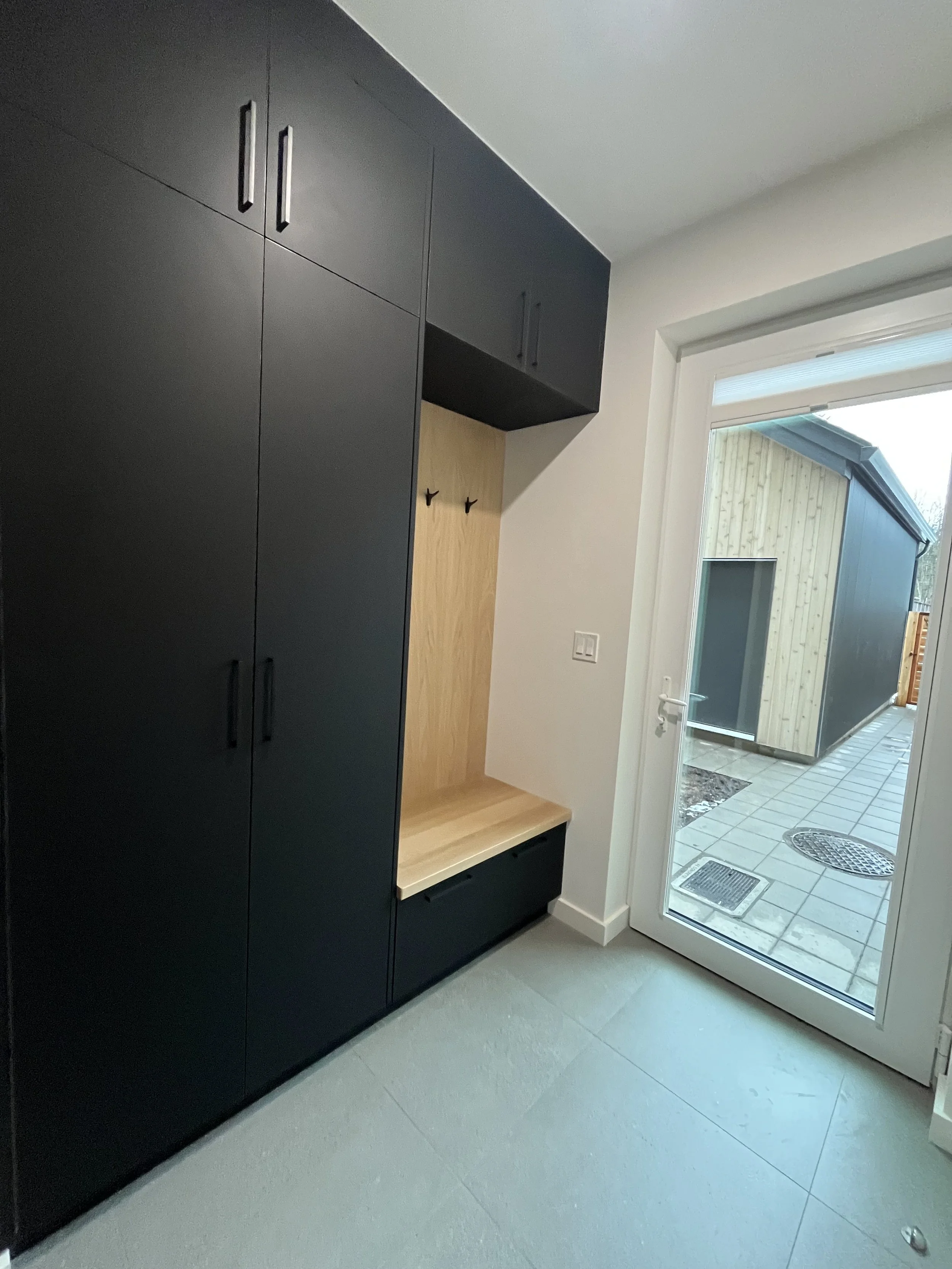 Interior view of an entryway with black built-in cabinets, a small wooden bench, and a glass door leading outside to a paved area with a building in the background.