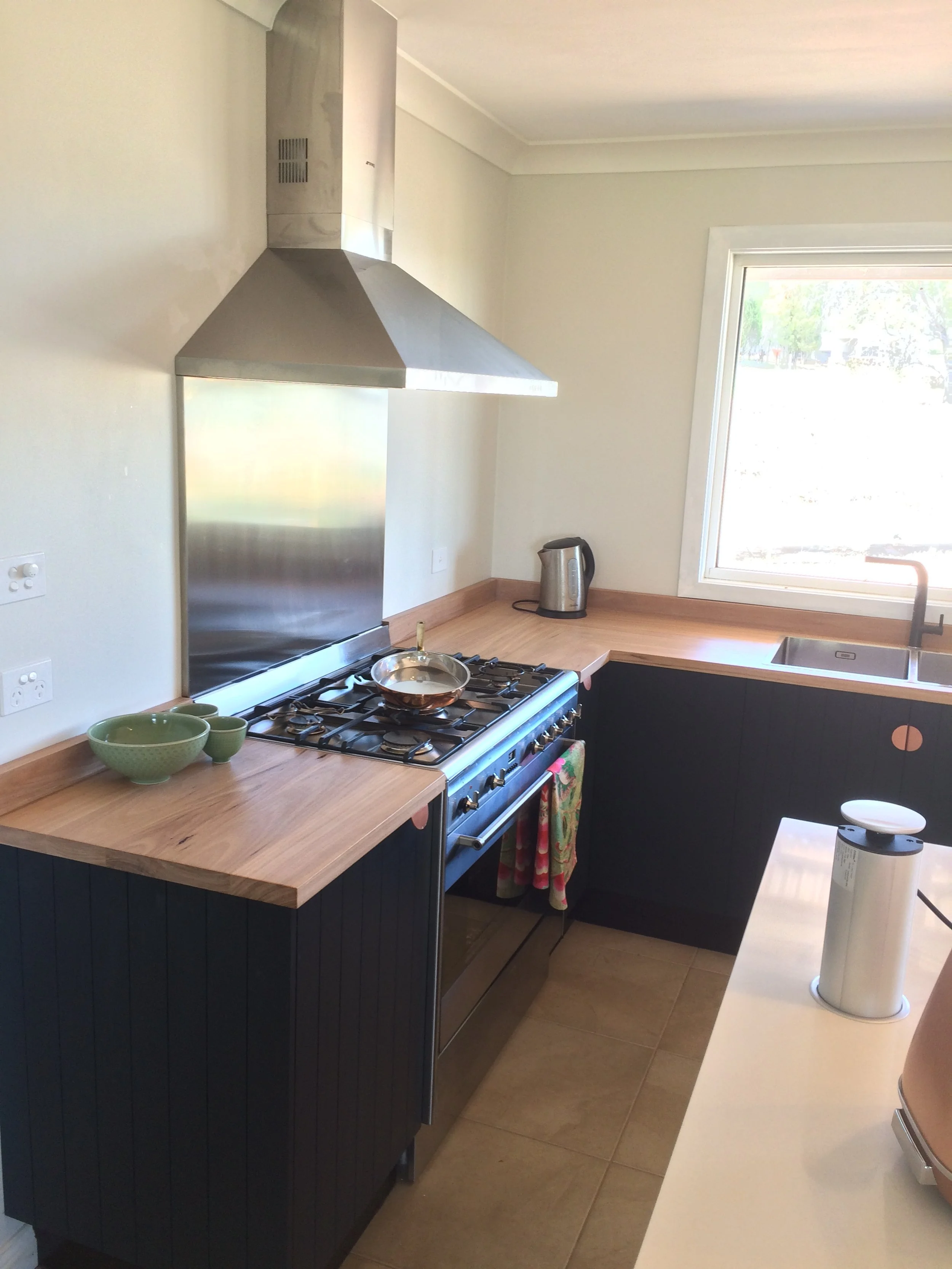 Kitchen with wooden countertops, a stainless steel range hood over a stove, a small skillet on the stove, a green bowl and cup on the counter, a kettle near the window, and a white countertop with a container on top.