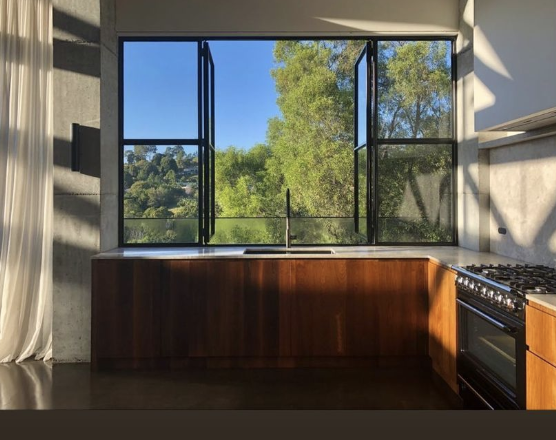 Kitchen with a large open window overlooking green trees and a clear blue sky, wooden kitchen cabinets, and a stove on the right.