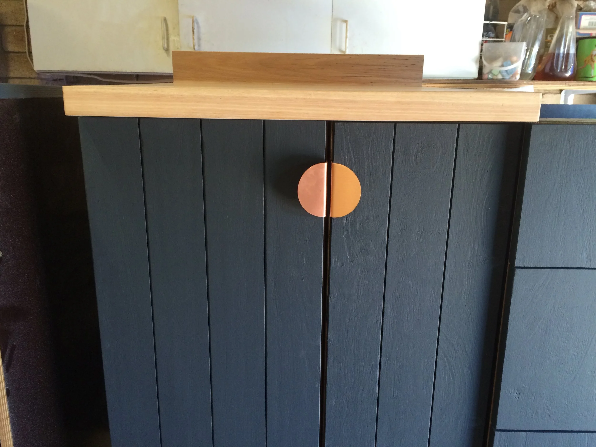 Close-up of a black cabinet with vertical wooden panels and a round copper-colored handle, topped with a light wood surface.