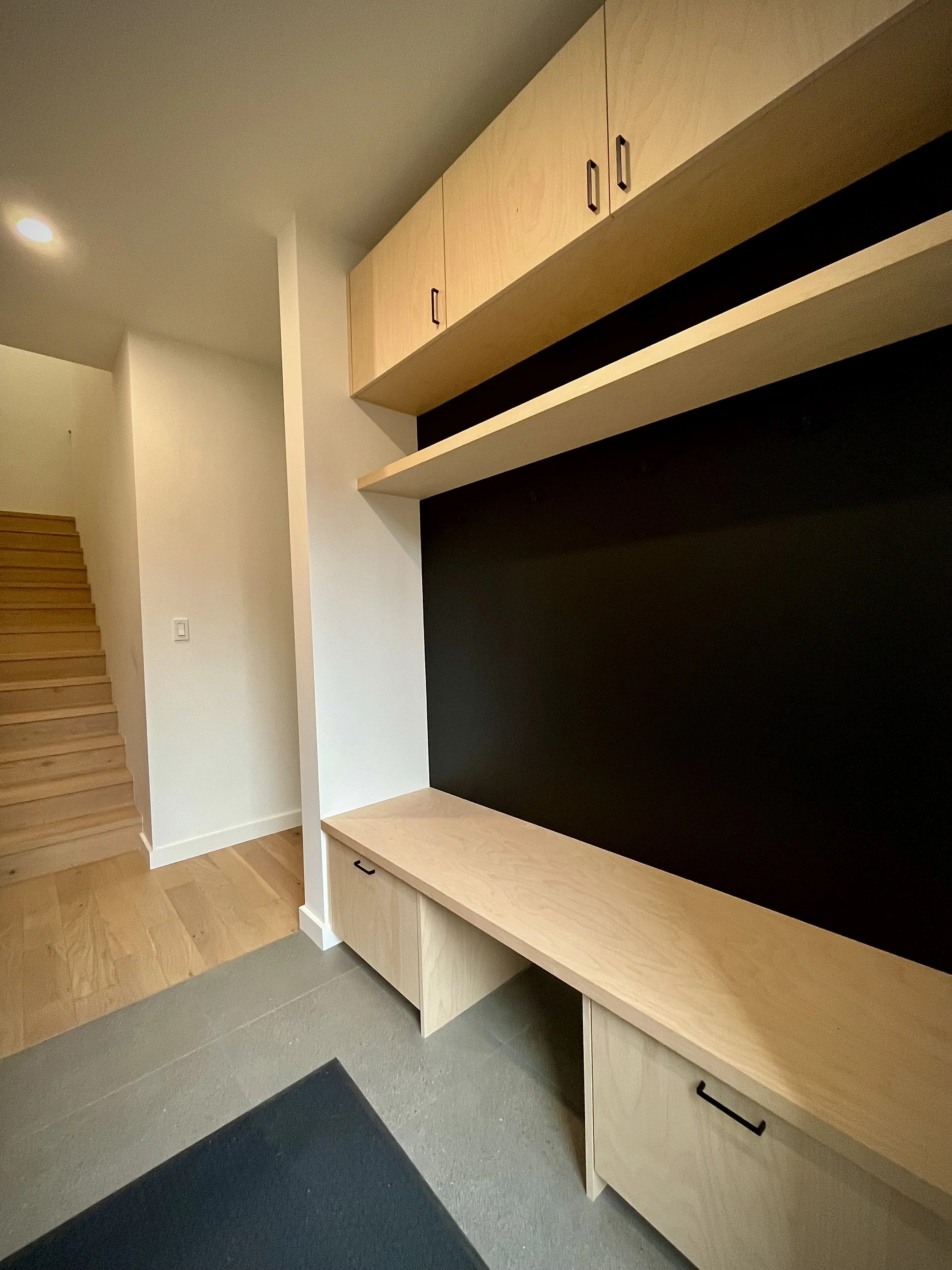 Interior of a modern house, showing a staircase with wooden steps to the left, a white wall, and a custom built-in wooden storage unit with upper cabinets, a shelf, and lower drawers against a black accent wall.