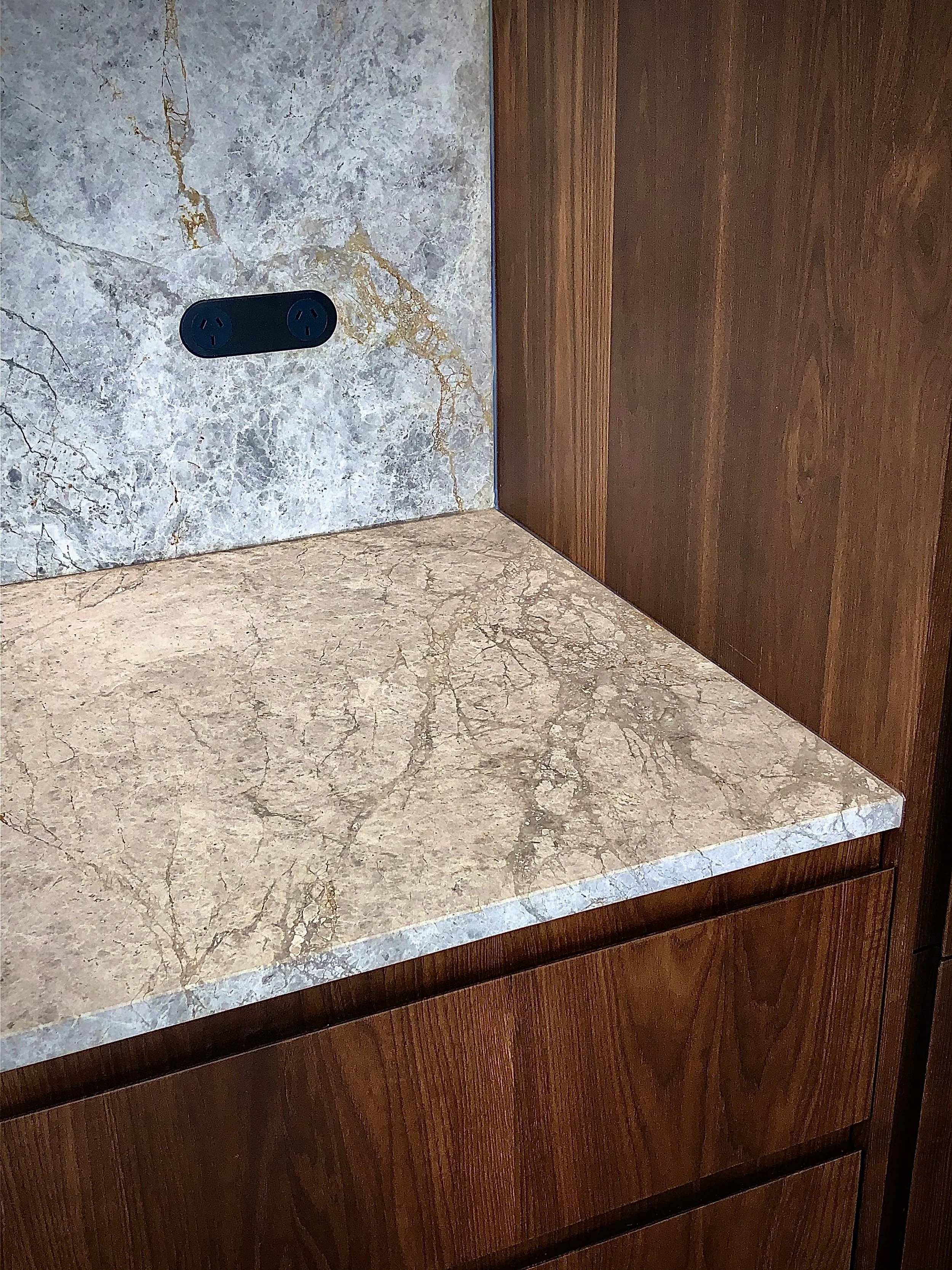 Close-up photo of a marble countertop with an electrical outlet on a marble wall and wooden cabinetry.