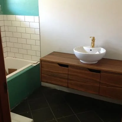 Bathroom with a wooden vanity, white vessel sink, gold faucet, and partial view of a shower with white subway tiles and green accents.