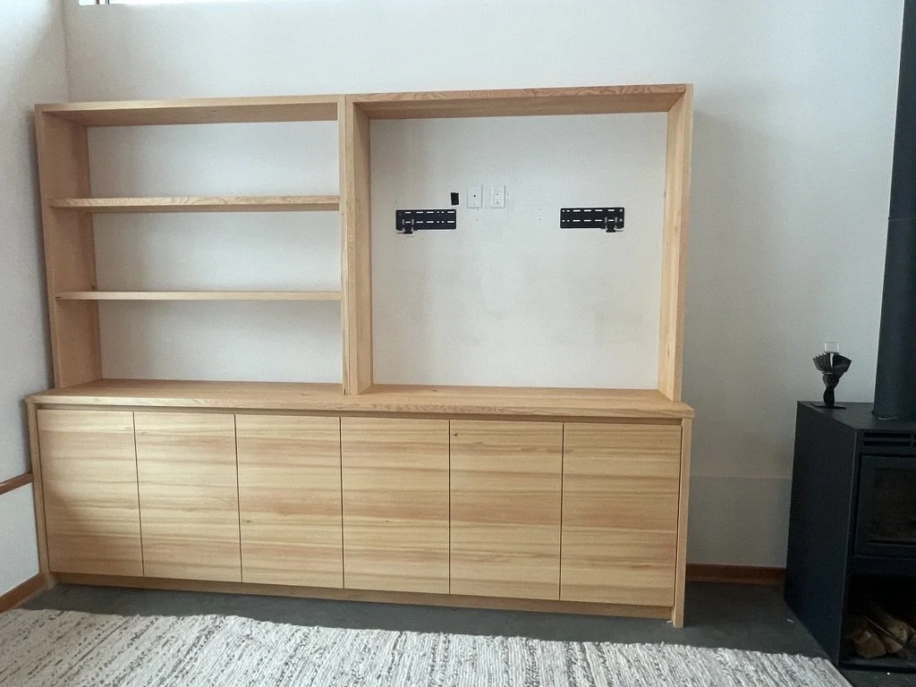 Unfinished wooden cabinet with shelves on the left and a large empty space on the right, situated against a white wall with visible outlets and brackets for a TV, next to a black wood stove.