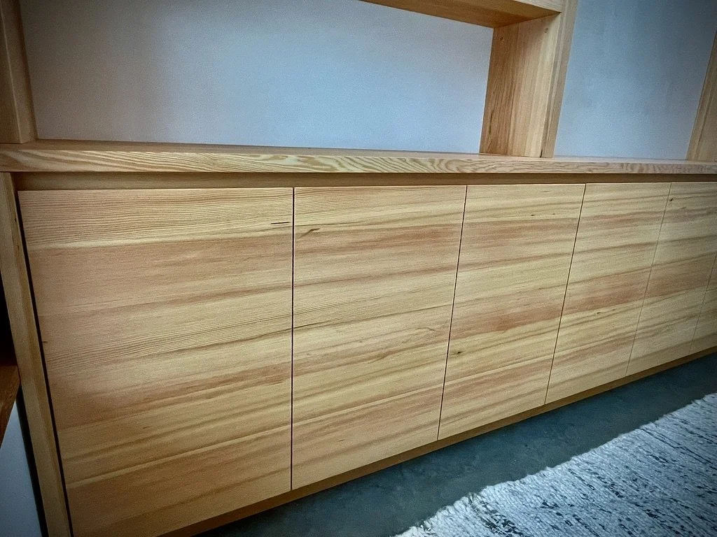 A wooden sideboard with six cabinet doors, placed against a light-colored wall, with a part of a shelf above and a rug on the floor.