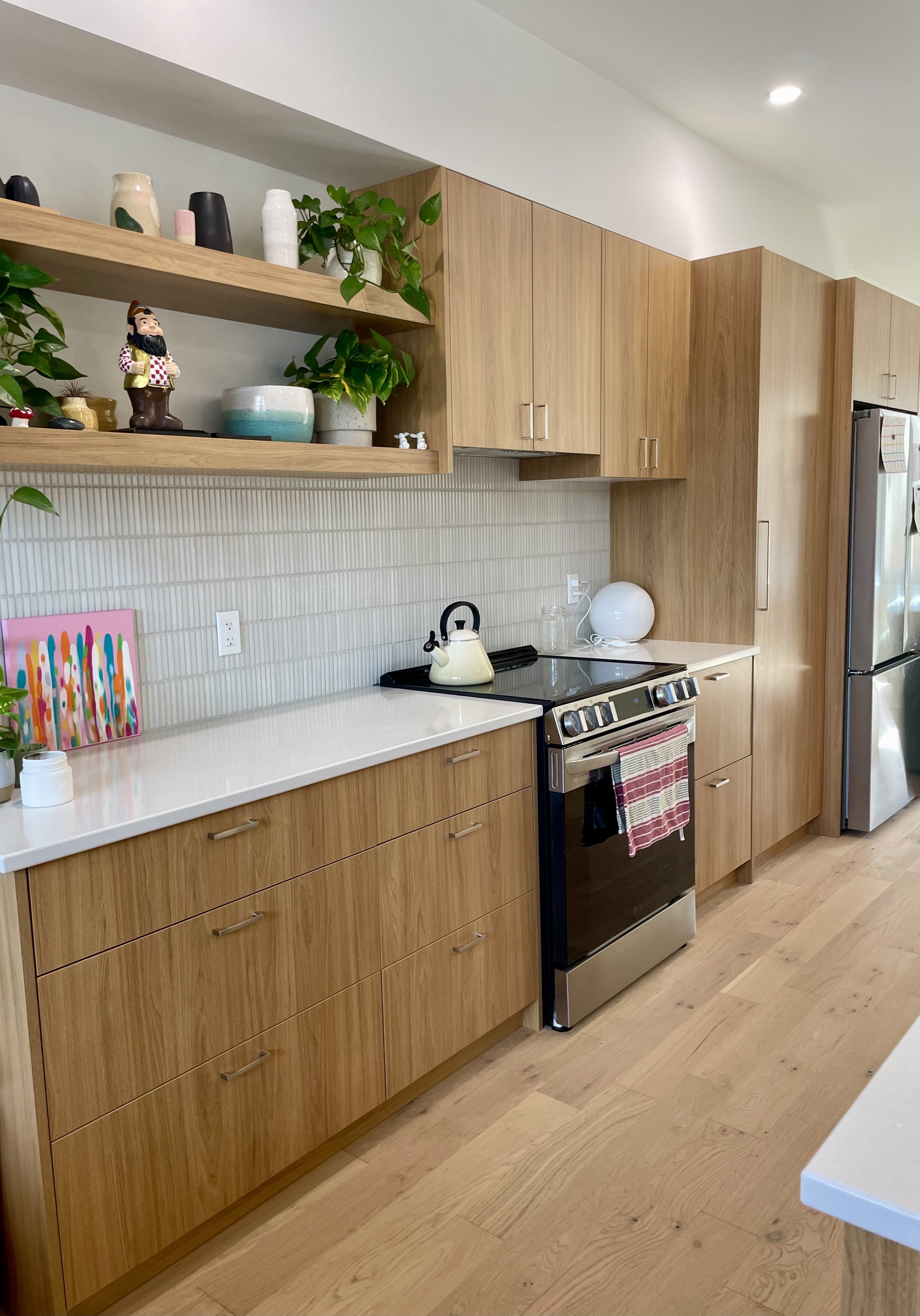 Modern kitchen with wood cabinets, white countertop, stove, kettle, potted plants, shelves with vases and decorative items, and a stainless steel refrigerator.