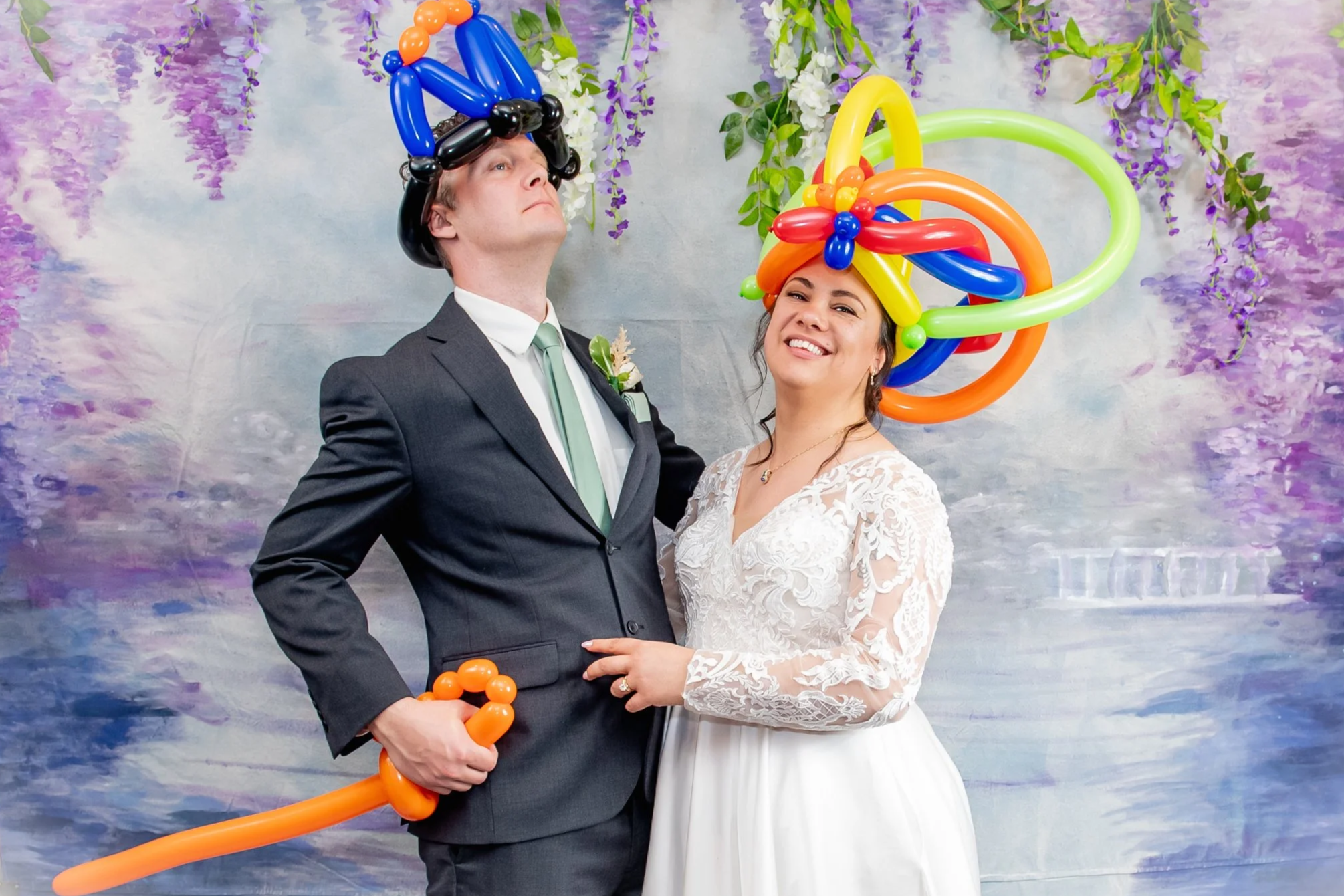 A bride and groom stand in front of a painted backdrop with purple flowers and greenery, both wearing colorful balloon hats. The groom wears a black suit and holds a balloon sword, while the bride is in a white wedding dress, smiling and holding the groom's hand.