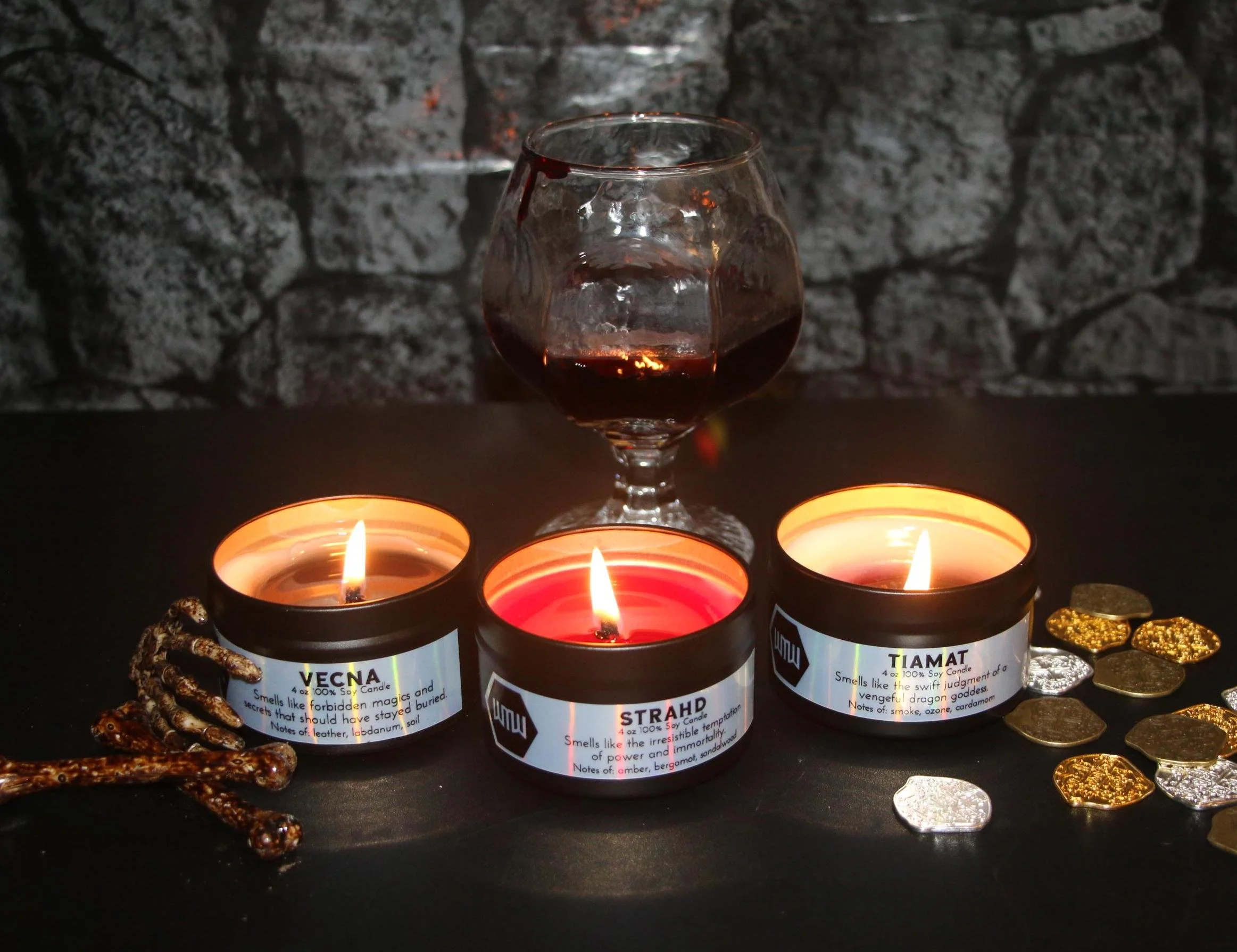 Vanity set with three lit candles in labeled holders, a glass of red wine, and scattered gold and silver coins on a dark surface against a dark brick wall background.
