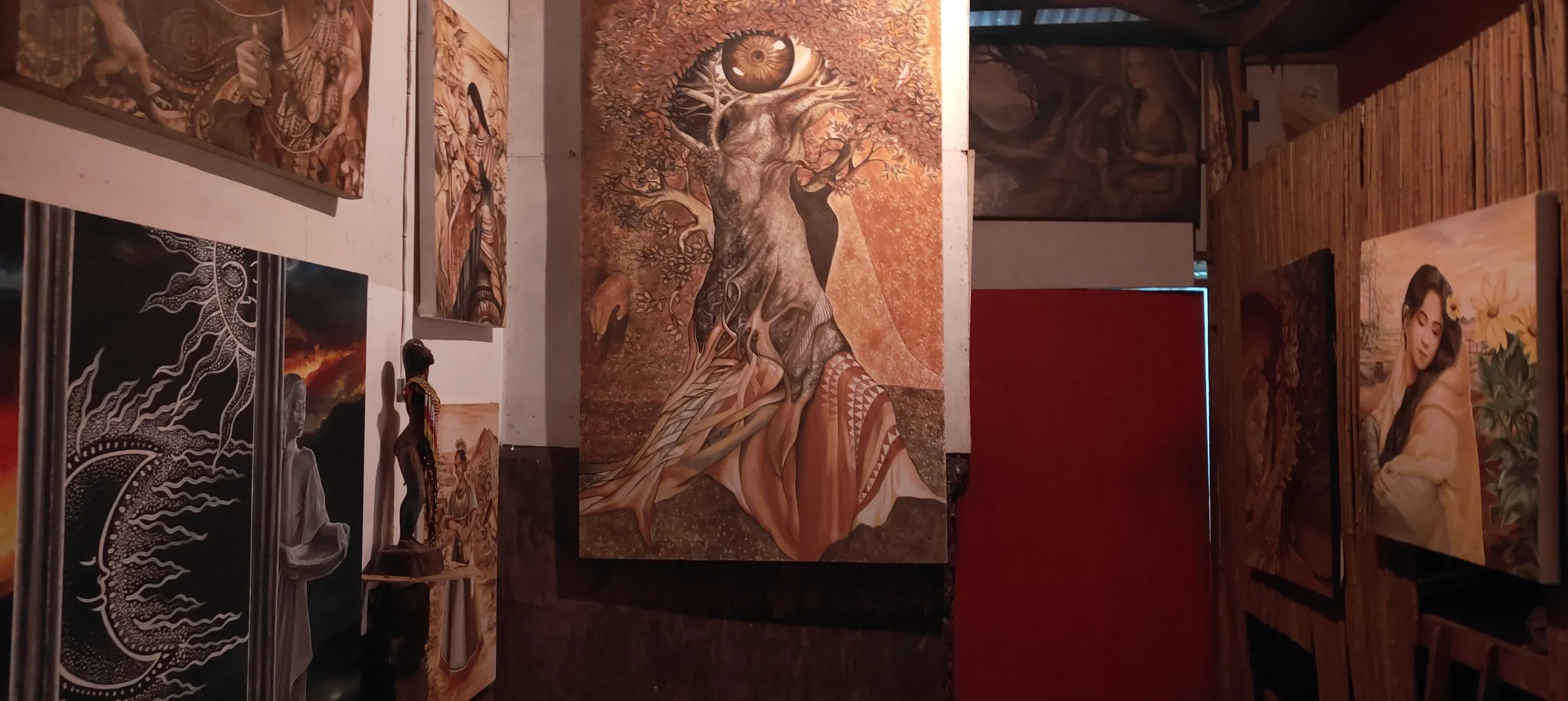 Art gallery with various paintings, including a large artwork of a tree with an eye and face in the center, and a sculptural figure of a woman among paintings.