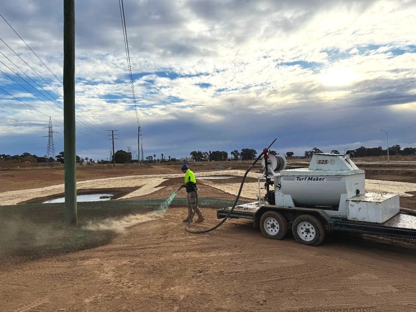 Worker operating turf-making equipment on a dirt construction site, under cloudy sky.