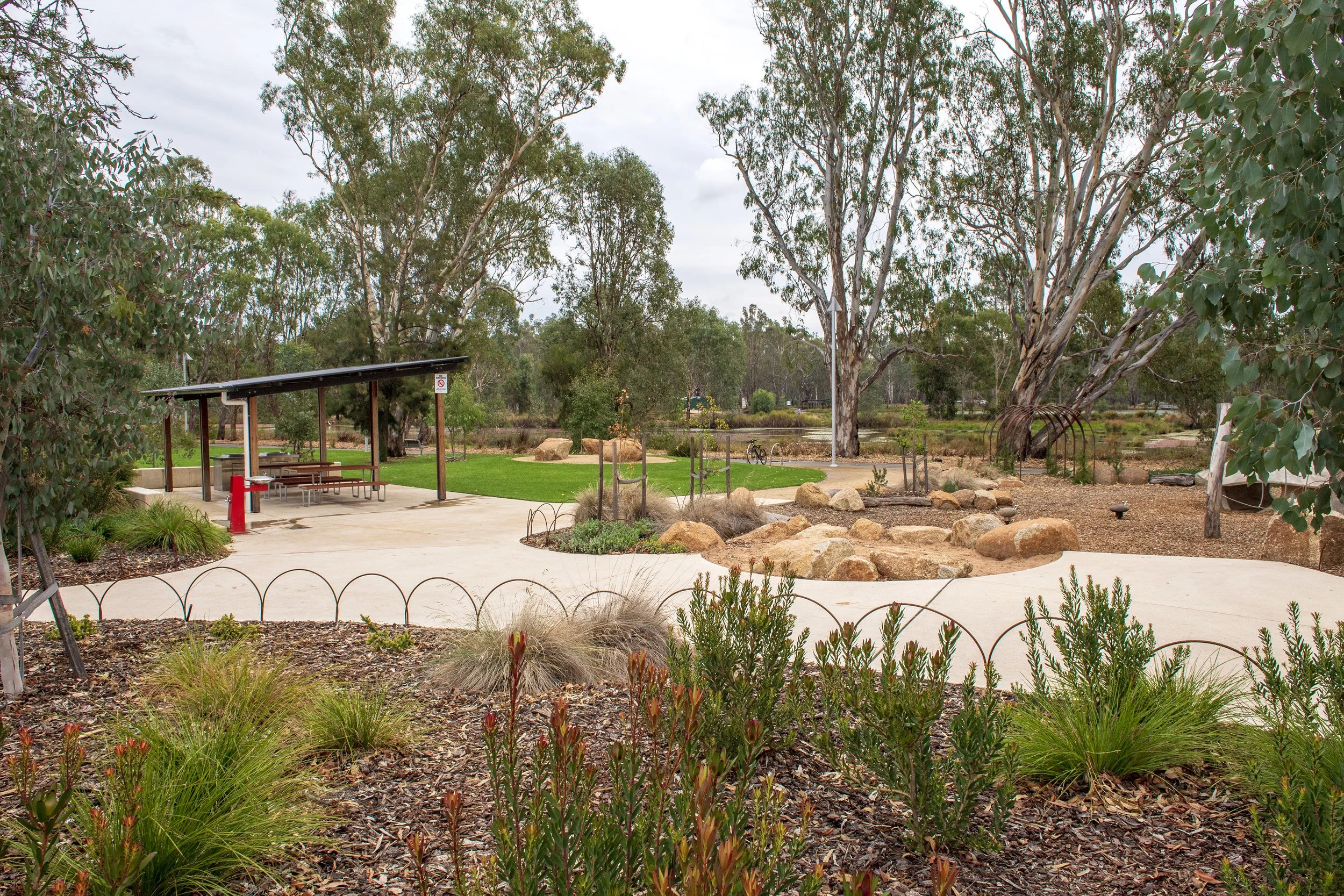 A park with a concrete pathway, a shelter with picnic tables, a fire hydrant, and large trees in the background. There are rocks and small plants along the pathway.