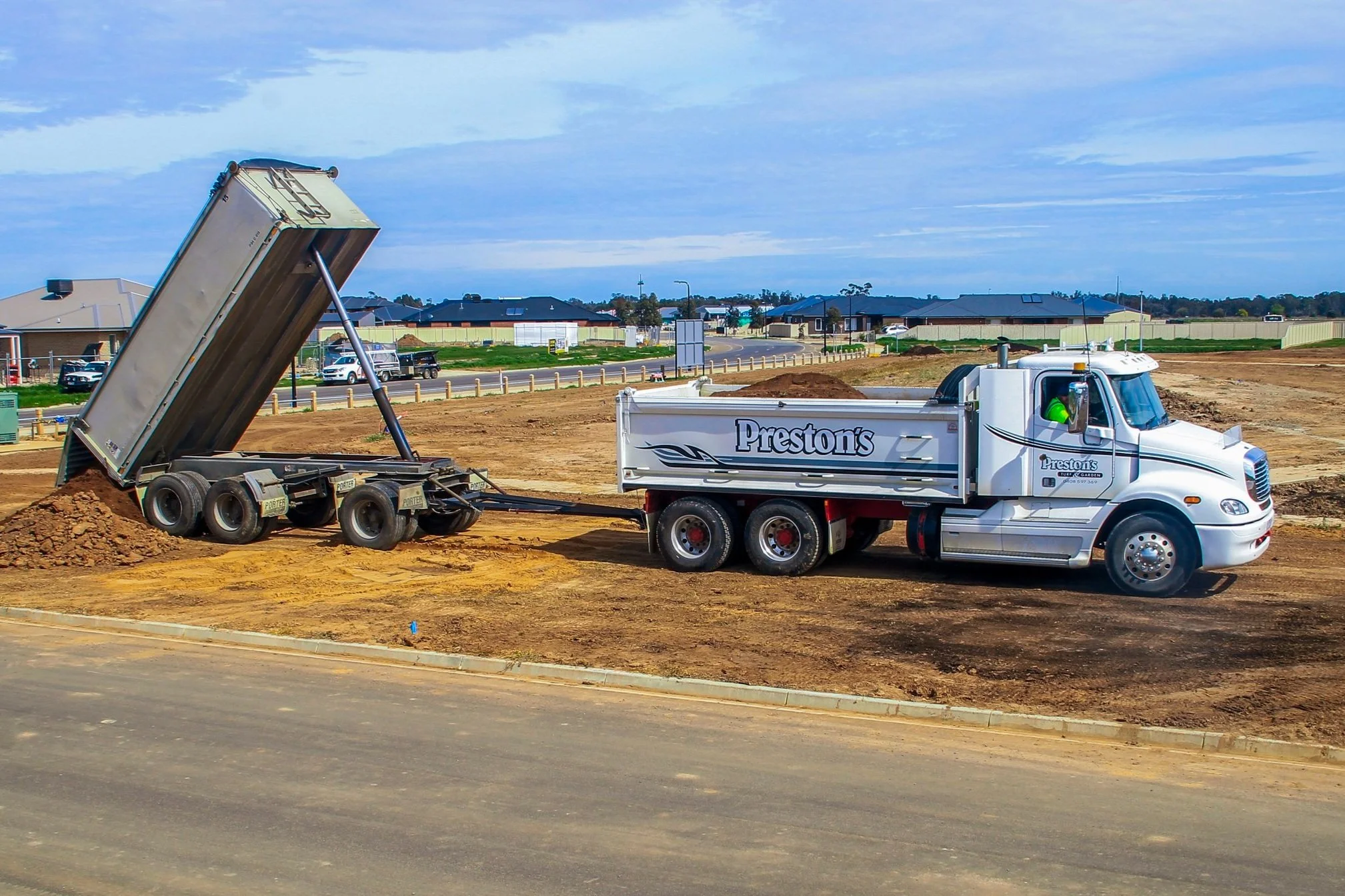 A construction site with a dump truck unloading dirt into another truck on a dirt lot, with houses and blue sky in the background.