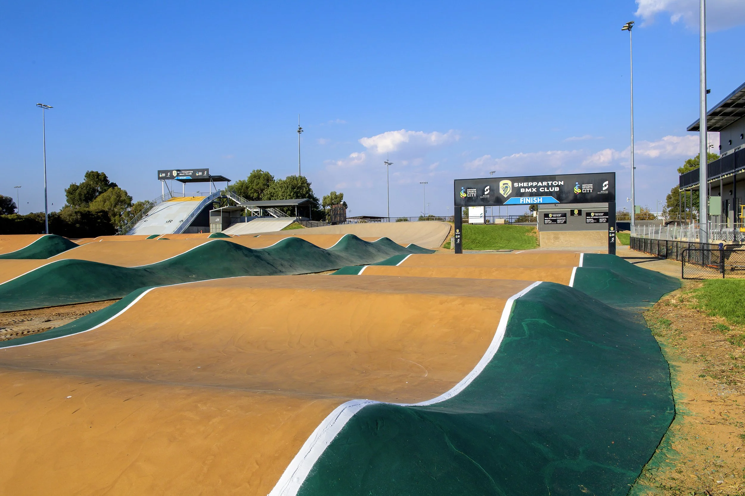 An outdoor BMX dirt jump track at Shepparton BMX Club with multiple dirt mounds, a viewing platform, and a finish line sign under a clear blue sky.
