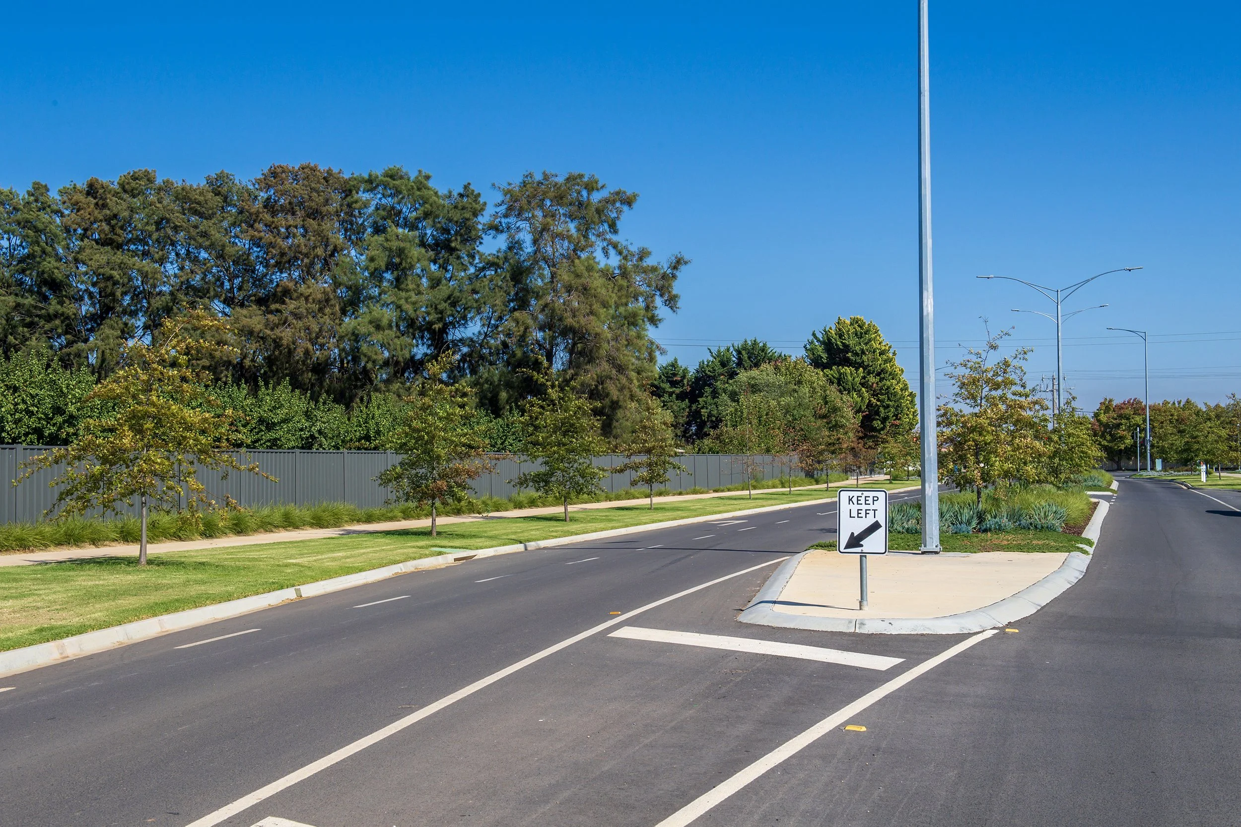 A street with trees, a sidewalk, and a tall streetlamp under a clear blue sky. A sign instructs to keep left, placed on a small median with landscaping.