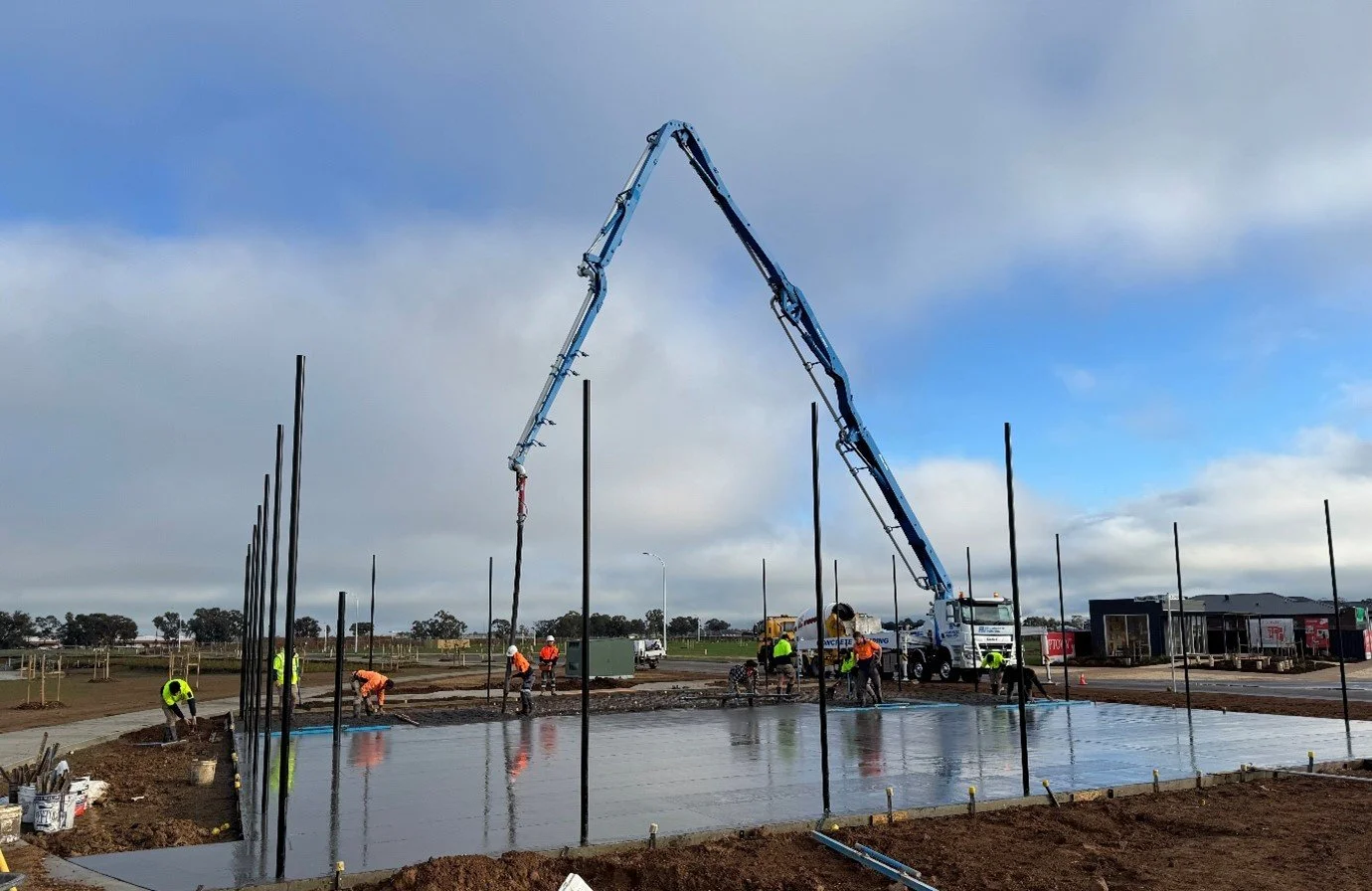 Construction workers pouring concrete on a foundation with a concrete pump truck and support poles around the area.
