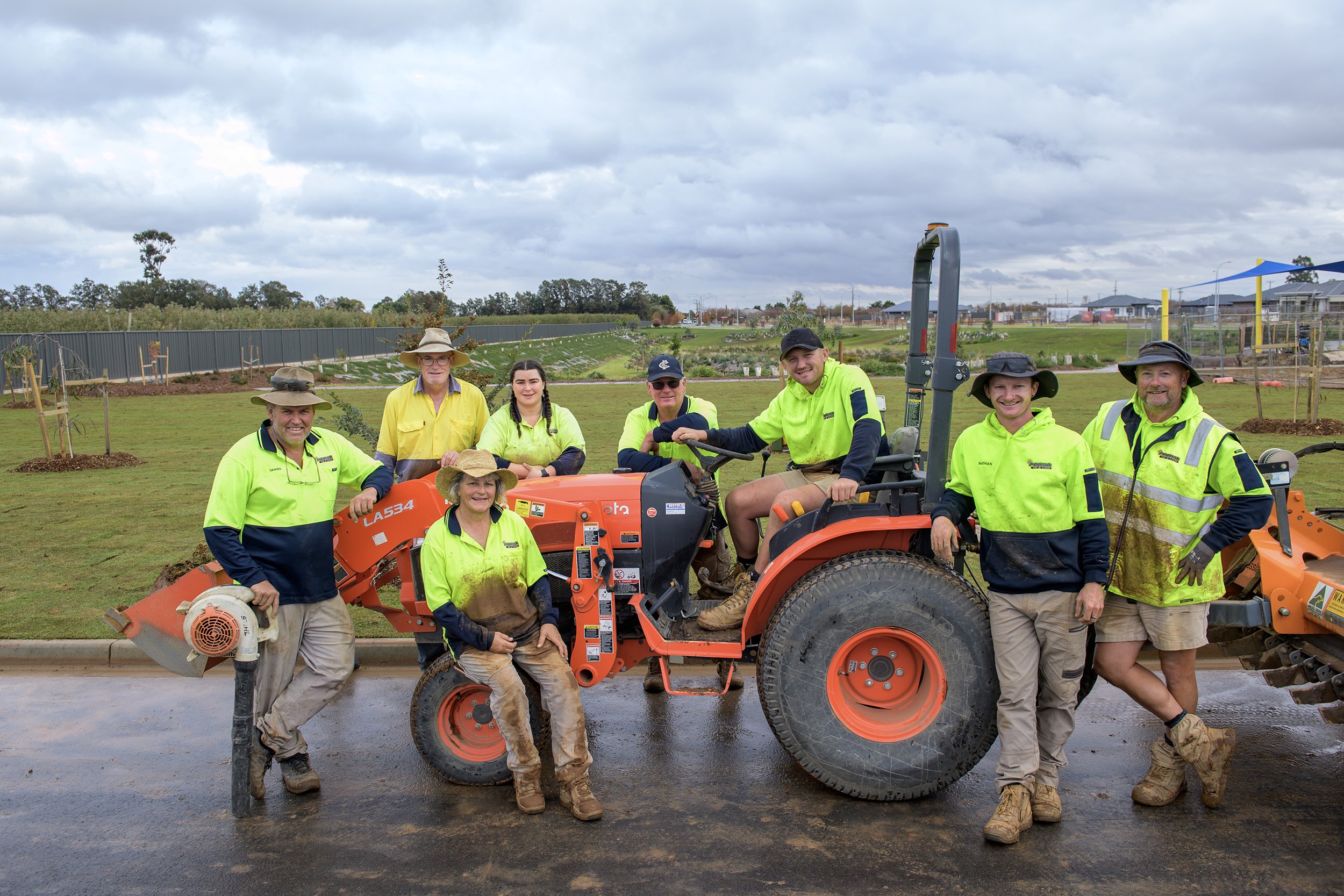 Group of people in high-visibility clothing on a construction site with a skid steer loader in the center, surrounded by grass and trees under a cloudy sky.
