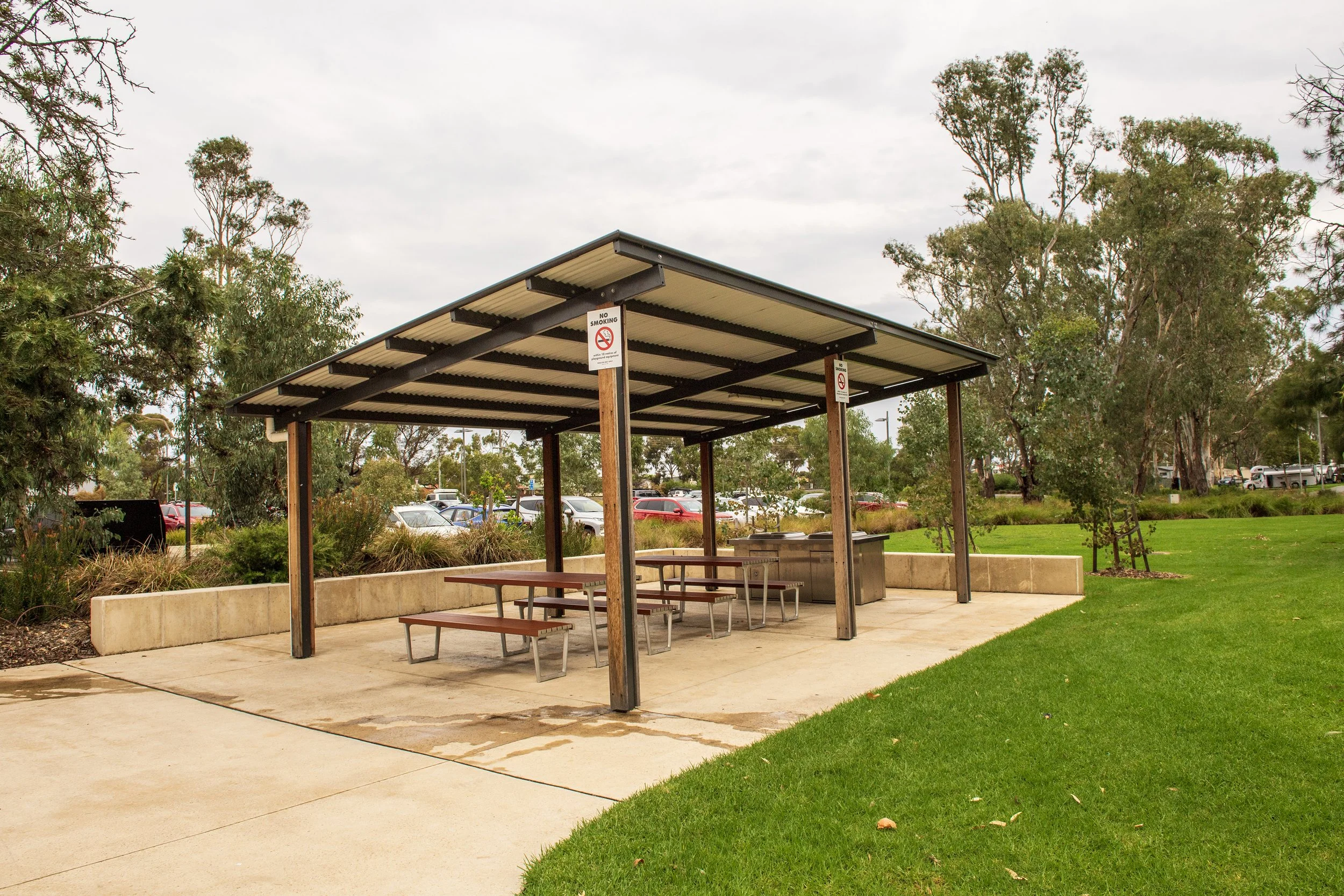 A picnic shelter with a metal roof and wooden supports in a park, surrounded by green grass and trees, with parking visible in the background.