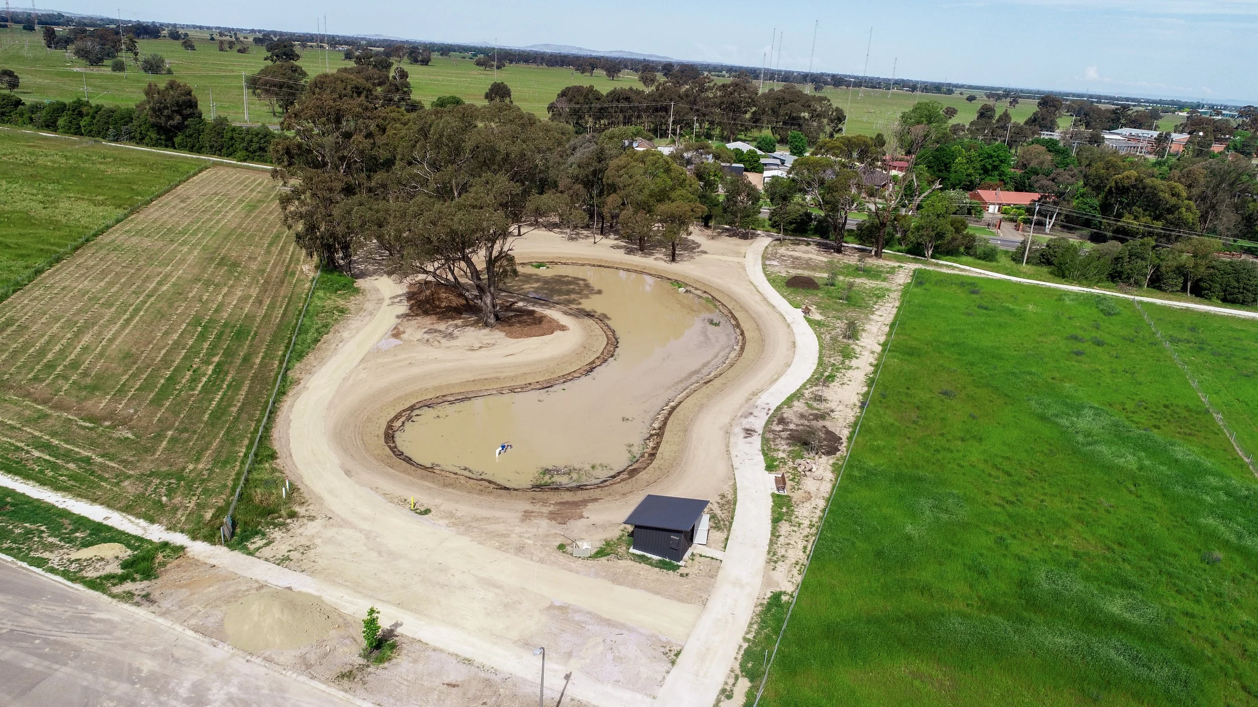 Aerial view of a horse track surrounded by green fields, trees, and a small black shed, with some water in the track.