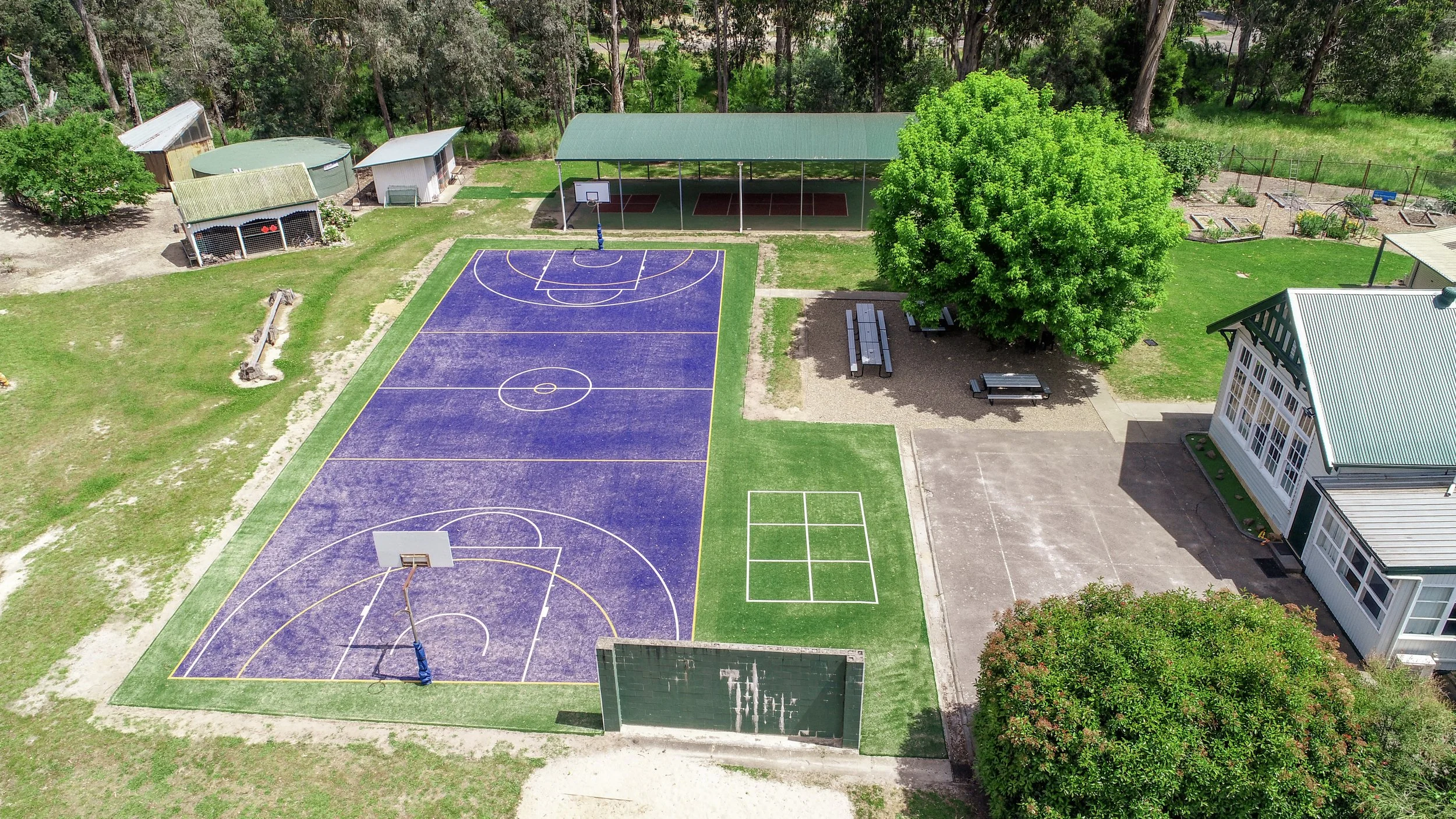 An aerial view of an outdoor sports area with a purple basketball court, a mini tennis court, a playground with a slide, and several surrounding trees and buildings.