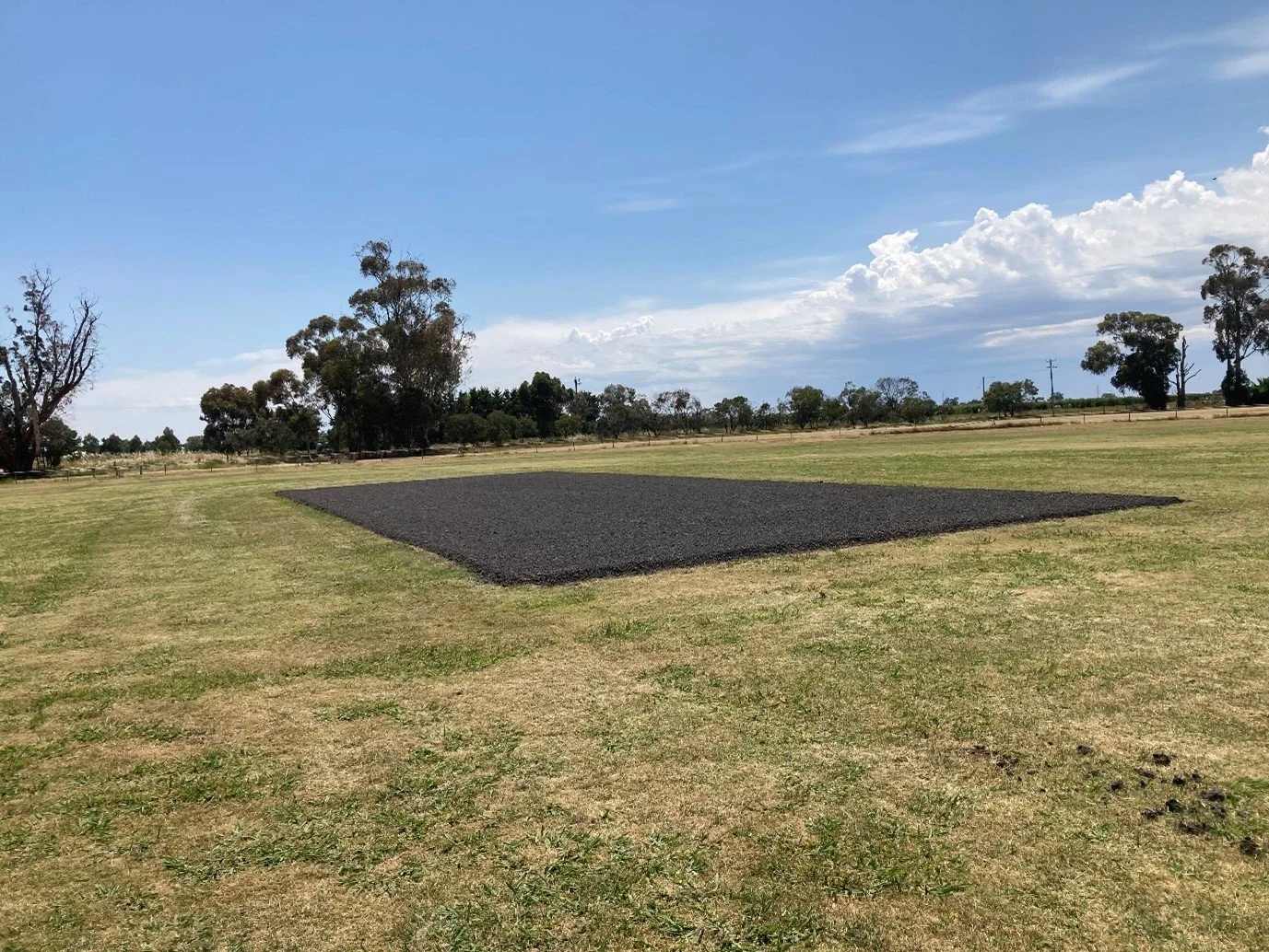 A rectangular patch of dark gravel on a grassy field with trees and blue sky in the background.