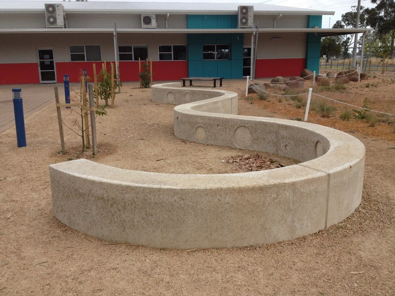 A circular concrete seating wall in a playground area outside a building with a red and teal facade, and small trees planted nearby.