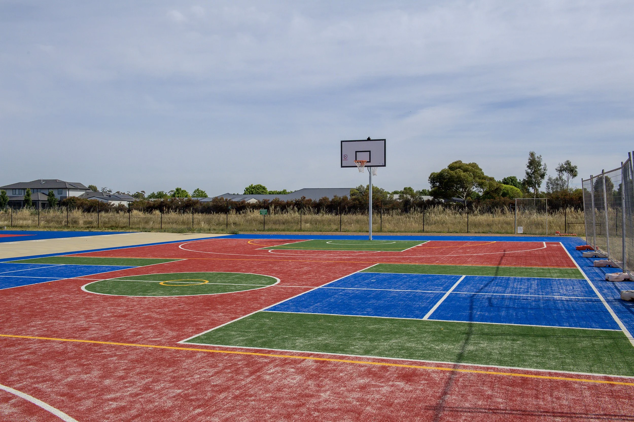 An outdoor multi-sport basketball court with colorful markings, fenced area, and a basketball hoop against a backdrop of trees and residential buildings under a cloudy sky.
