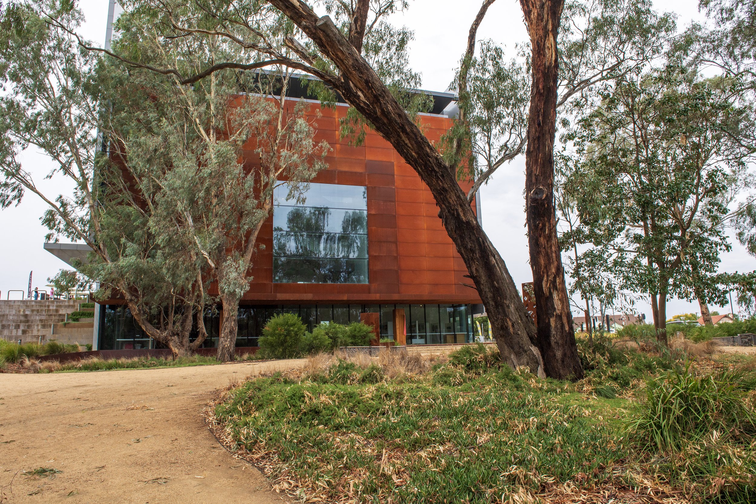 A modern building with a rust-colored exterior and large glass windows, surrounded by trees and a dirt pathway in a natural setting.