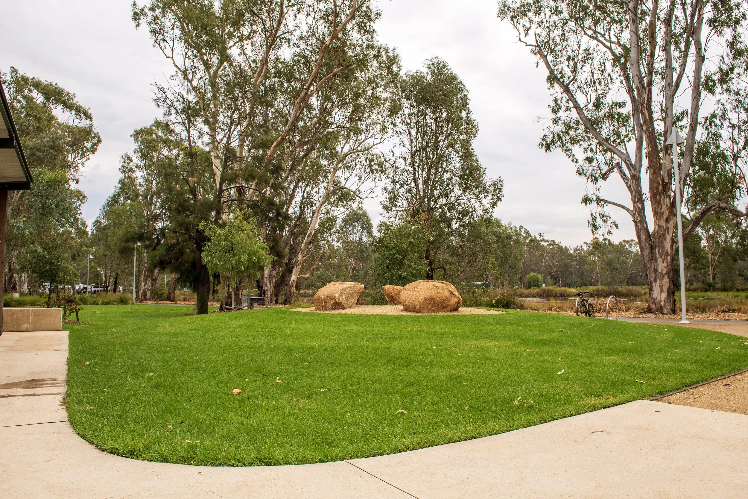 A park with a green grassy area, large trees, and three rocks in the background. Sidewalks and a bicycle rack are visible.