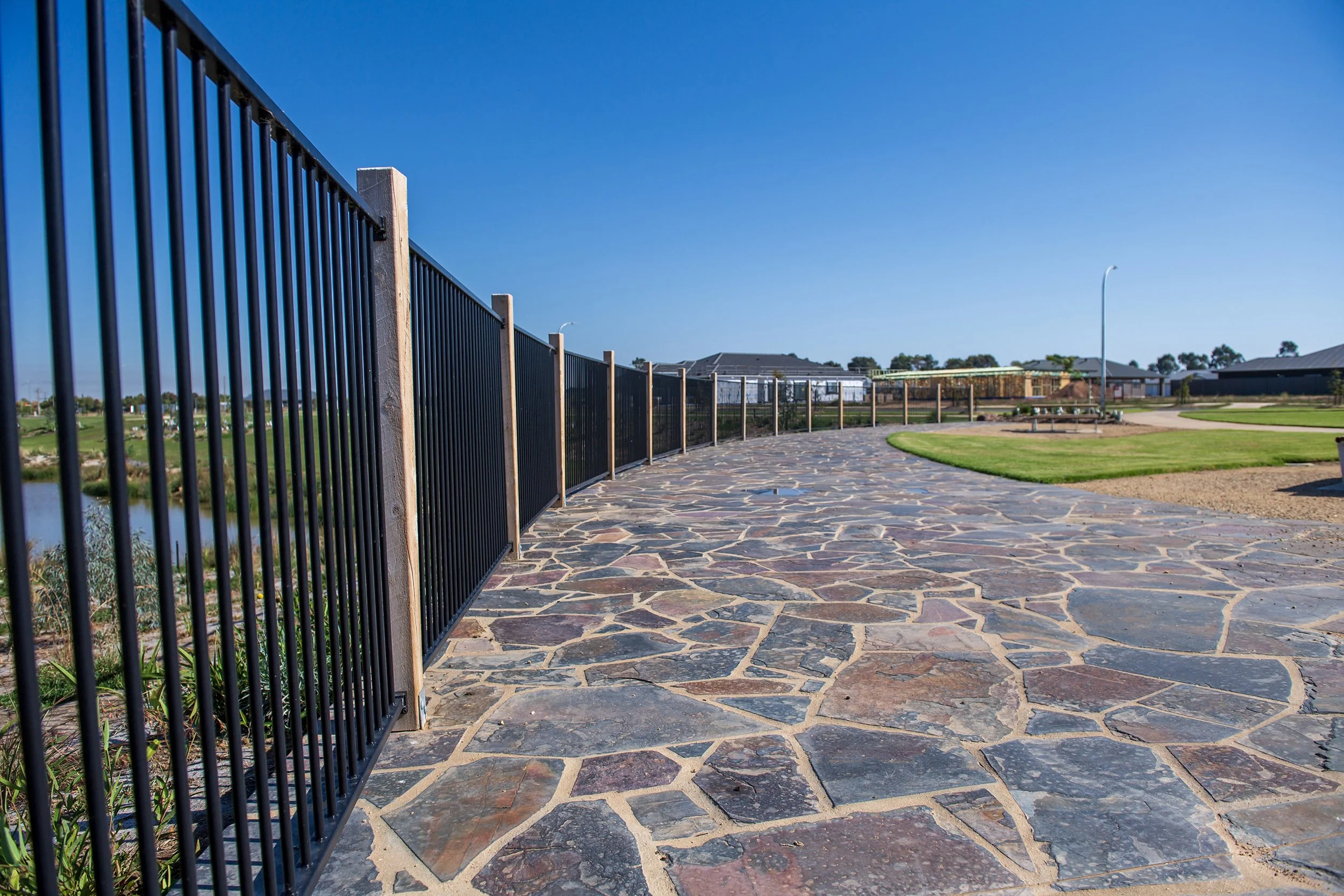 View of a paved stone walkway next to a black metal fence in a park with green grass and trees under a clear blue sky.