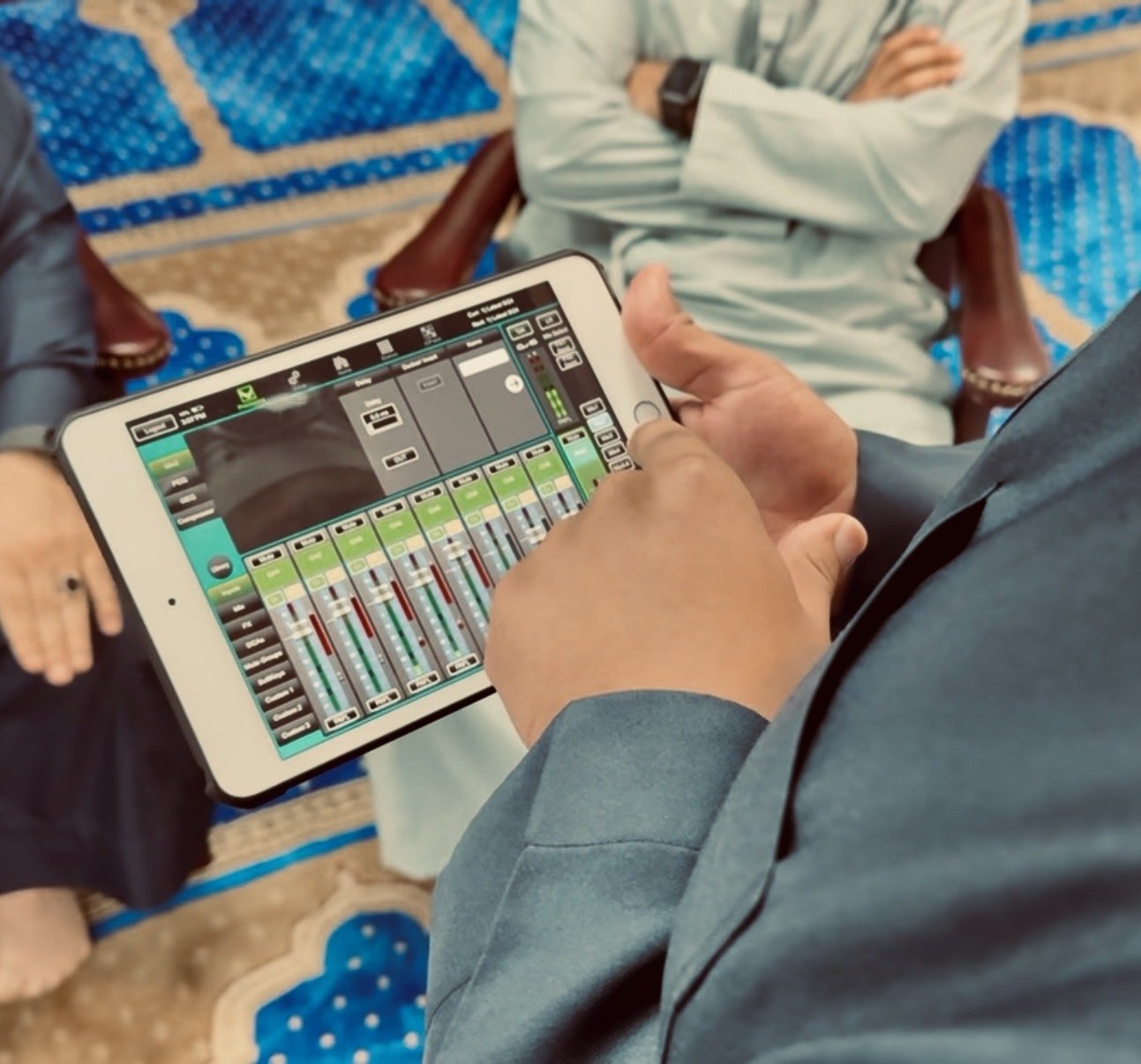 Person holding a tablet with a digital audio mixing console displayed on the screen, seated with two other people in a room with patterned carpet and traditional chairs.