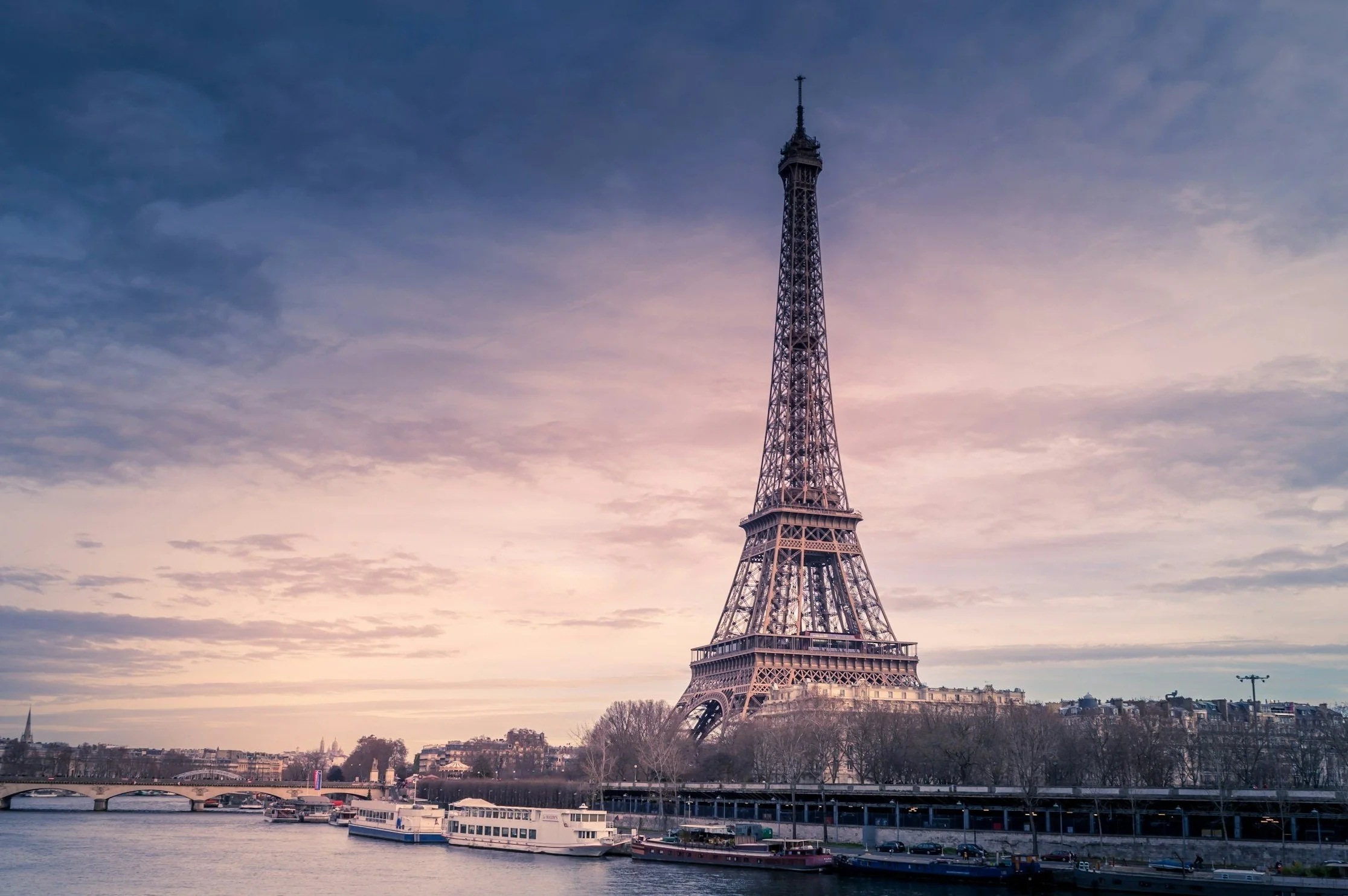 Eiffel Tower in Paris over the Seine River during sunset with boats and a cloudy sky.