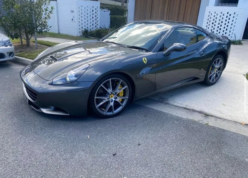 A dark gray Ferrari sports car parked on a residential street next to a driveway and lawn.