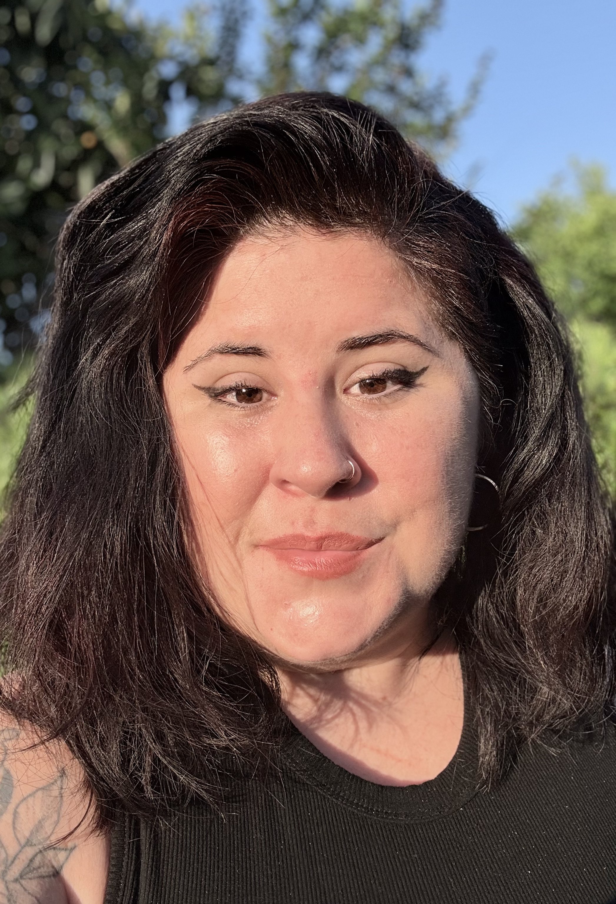 A woman with dark features and pale skin, smiling. She also has a nose piercing and a winged eyeliner look.