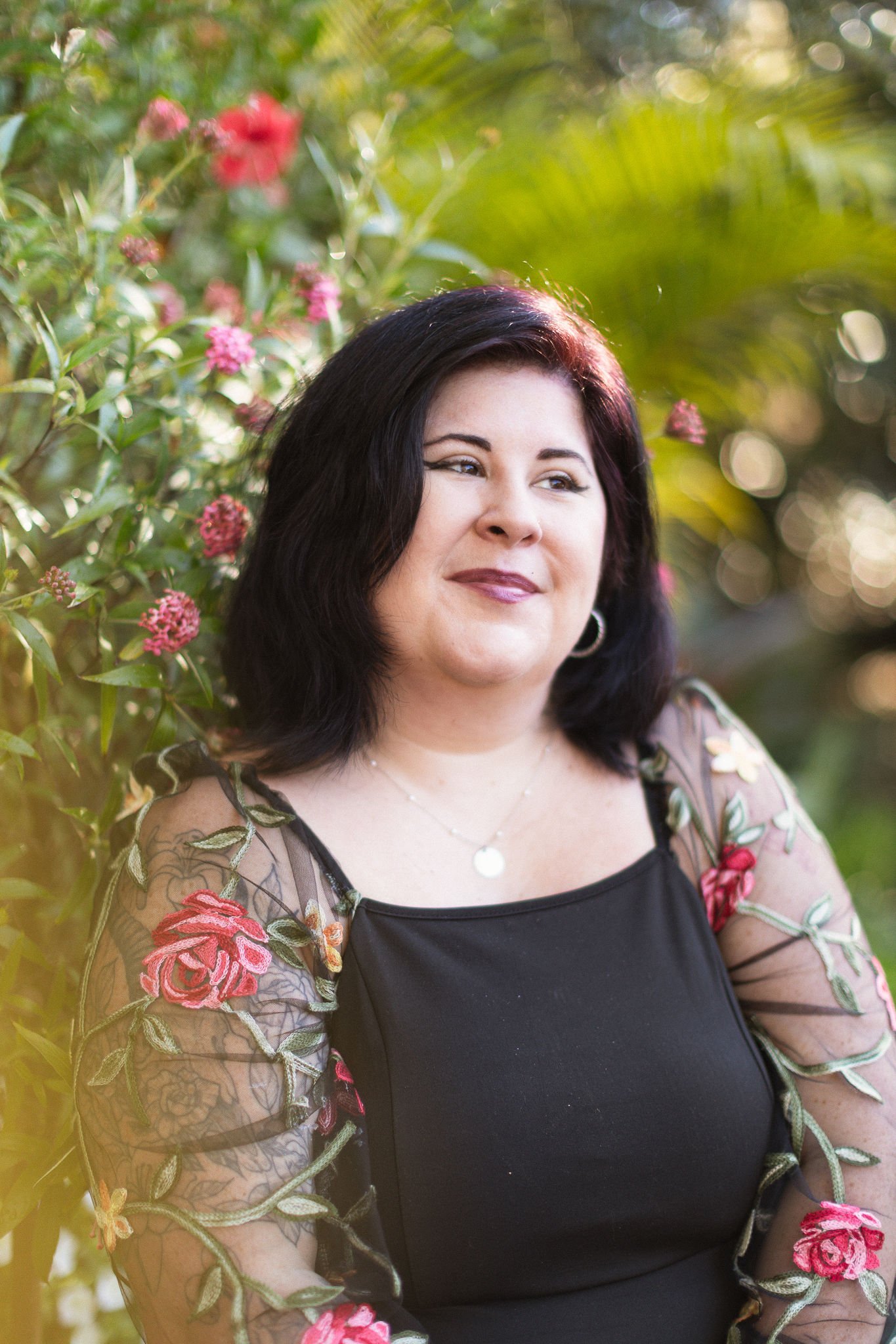 A woman with dark hair, wearing a black top and a sheer embroidered floral jacket, standing outdoors near pink flowers and green foliage.
